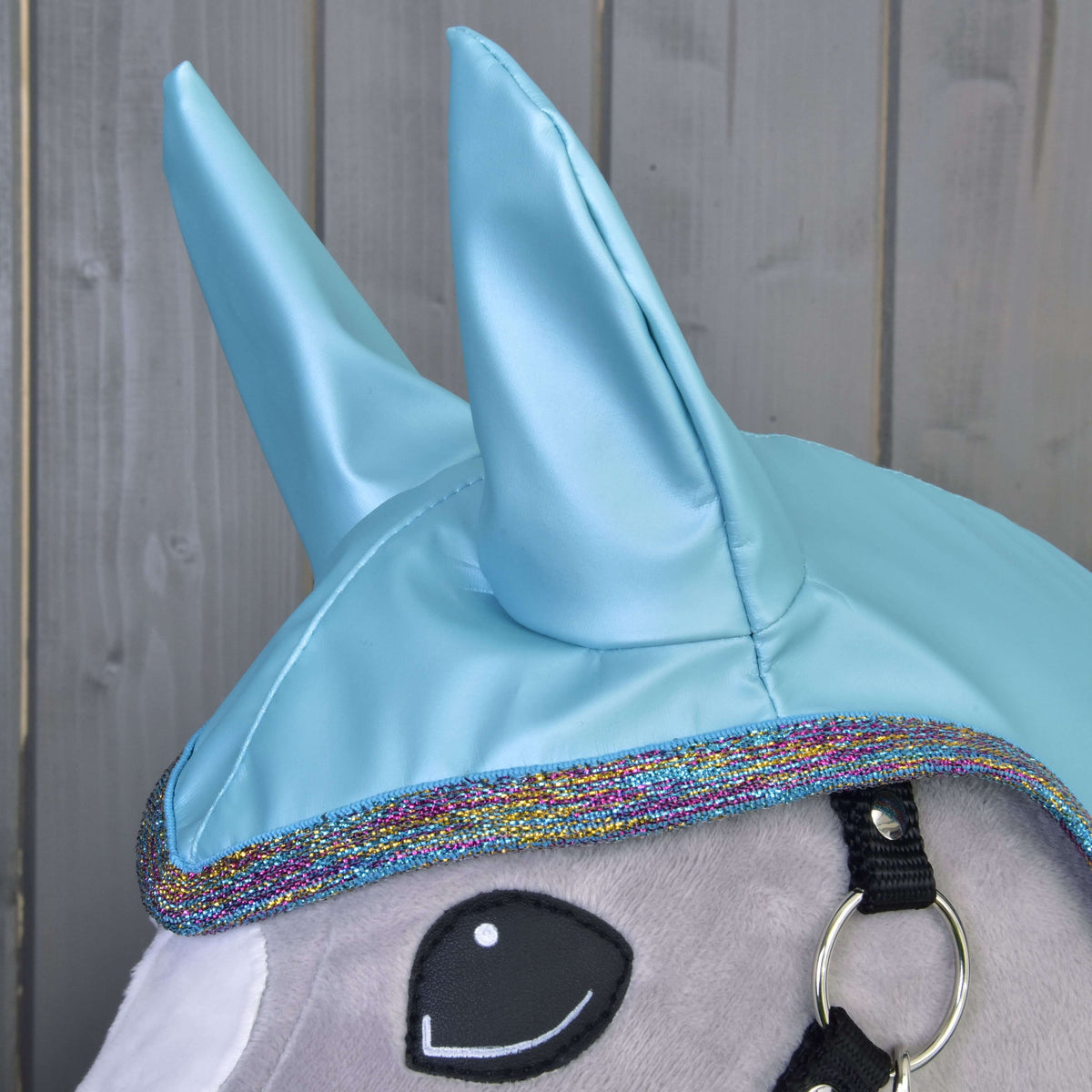 Detail Pláštenka Light Blue od Hobby Horse LarDen: plyšová hlava jednorožca s lesklými modrými rohmi, okrajom s dúhovým lemovaním, čiernou uzdečkou a vyšívaným okom v kreslenom štýle – ideálny pre hru za každého počasia na pozadí dreveného obloženia.