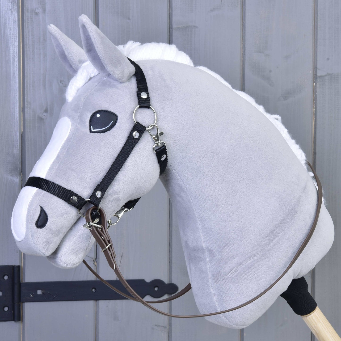 Sivý Hobby Horse od Hobby Horse LarDen s bielou hrivou, čiernou bezzubadlovou uzdečkou a svetlohnedými opratami, nasadený na drevenej tyči pred sivým dreveným plotom.