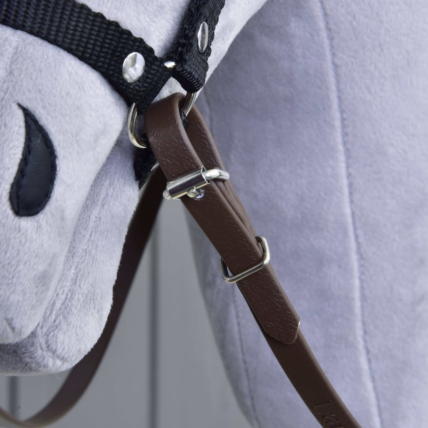 Detail Oprát Light brown od Hobby Horse LarDen na sivej plyšovej hračke koňa, zobrazujúci kožený remienok s prackou, čierne látkové opraty a uzdečku a vyšívané čierne oko.