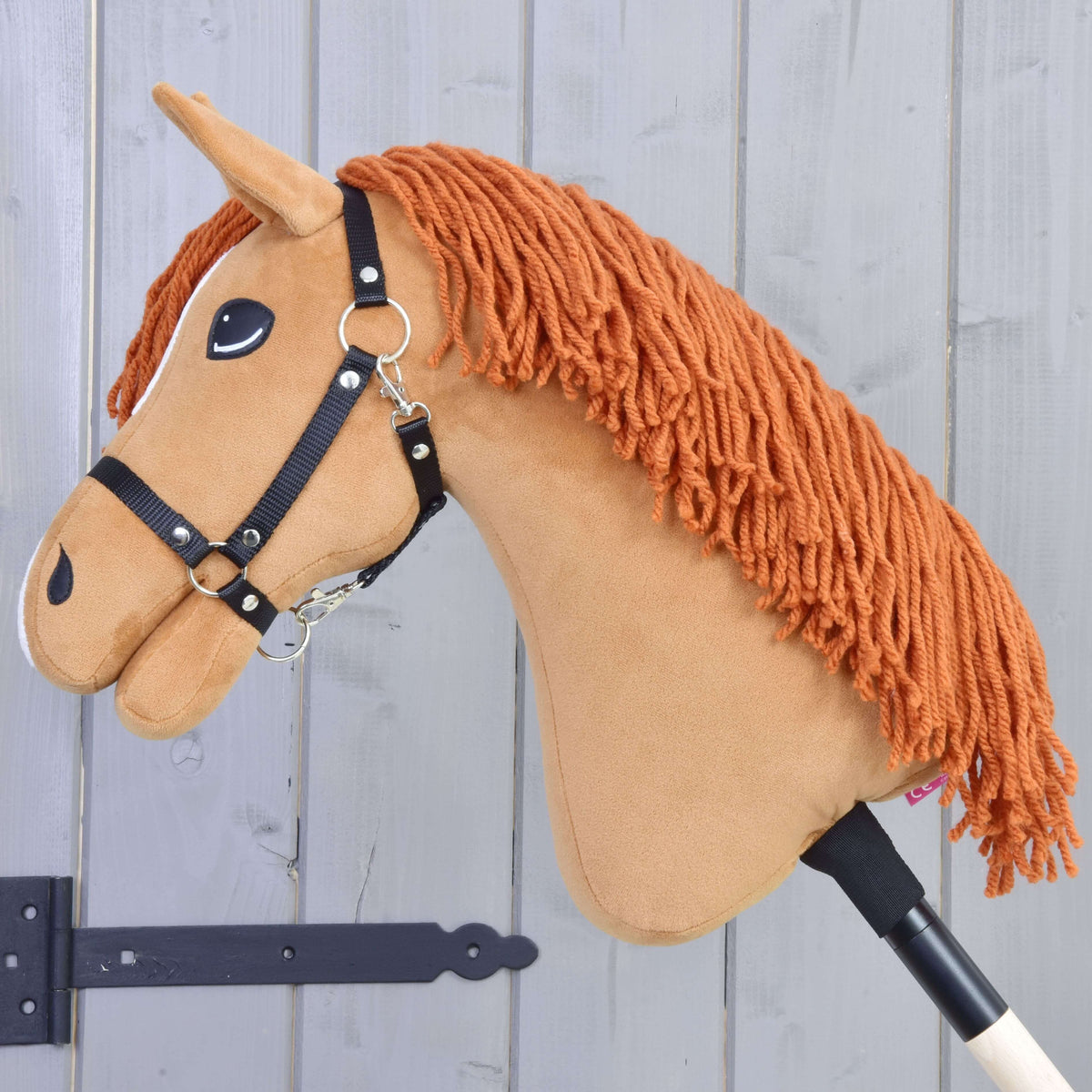 Hobby Horse Little Angel s čiernou ohlávkou od Hobby Horse LarDen má biele akcenty a oranžovú hrivu z priadze, je upevnený na tyči a zobrazený na svetlosivom drevenom pozadí.