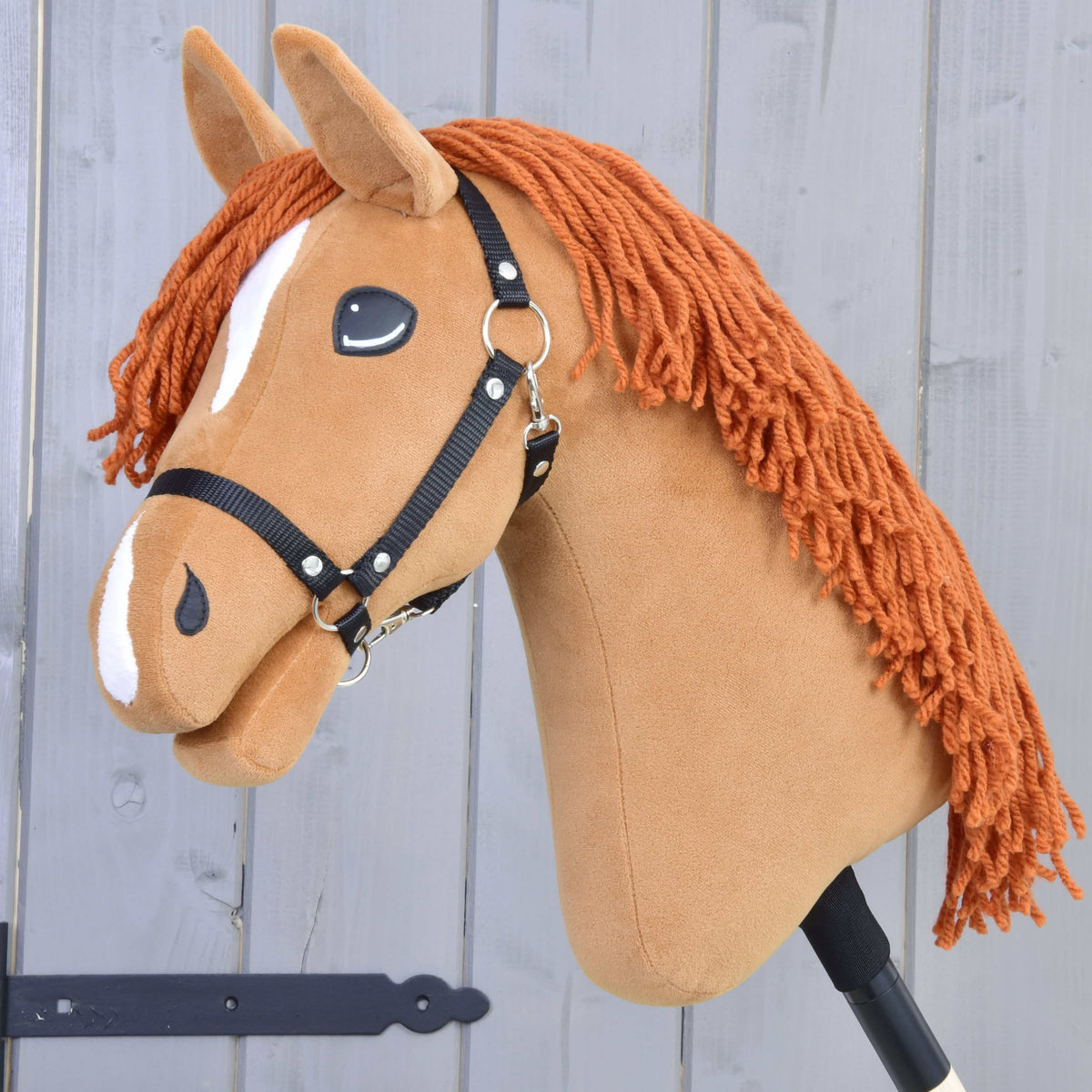Hobby Horse Little Angel s čiernou ohlávkou od Hobby Horse LarDen má hnedú plyšovú hlavu, oranžovú hrivu z priadze, biele znaky, čiernu ohlávku a je zobrazený na tyči na svetlosivom drevenom pozadí.
