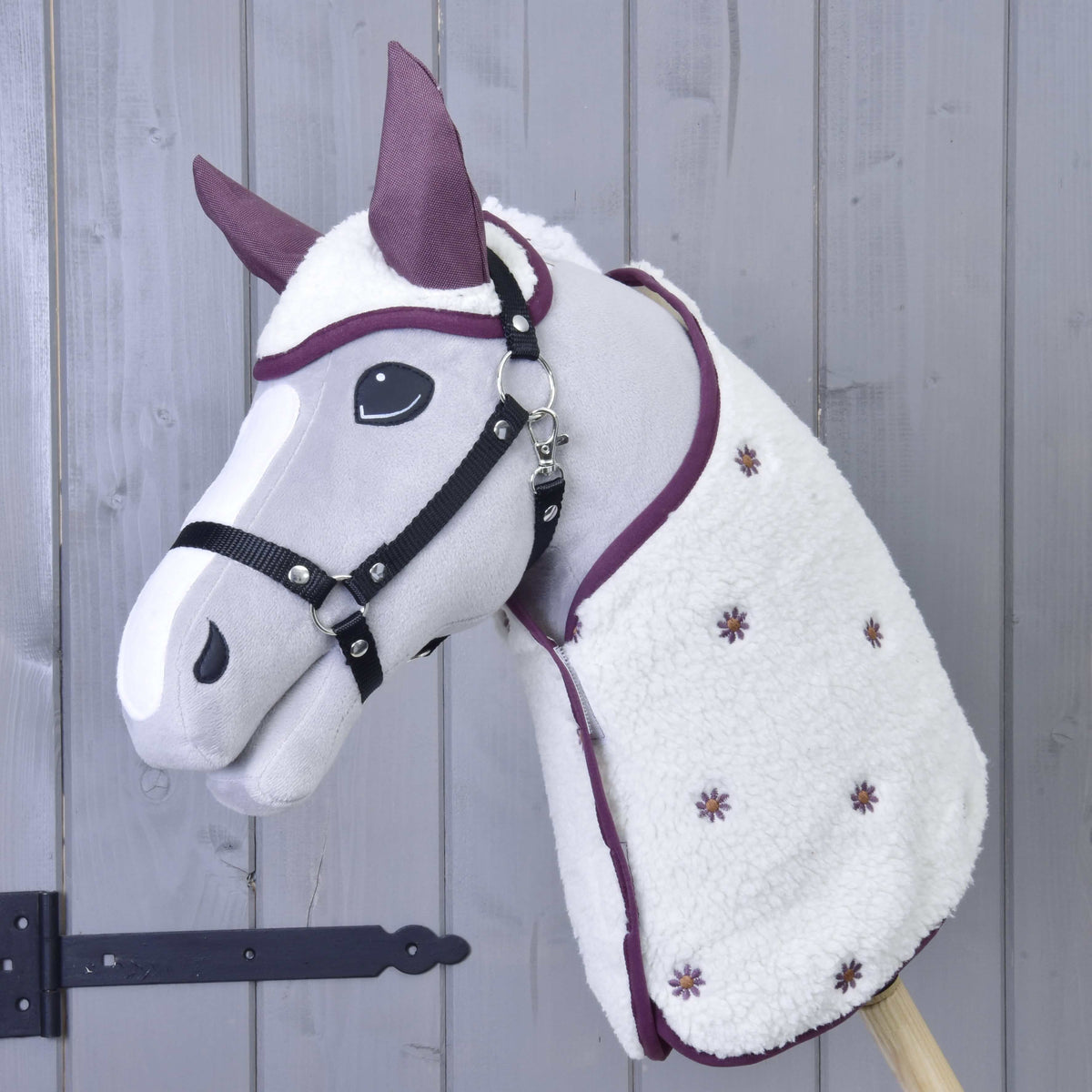 Set Loret od Hobby Horse LarDen má plyšového Hobby Horse so sivou tvárou, čiernou uzdečkou, fialovými detailmi, čabrakou a bielou dekou s drobnými fialovými kvetmi, naaranžovaného pred stenou z drevených panelov.