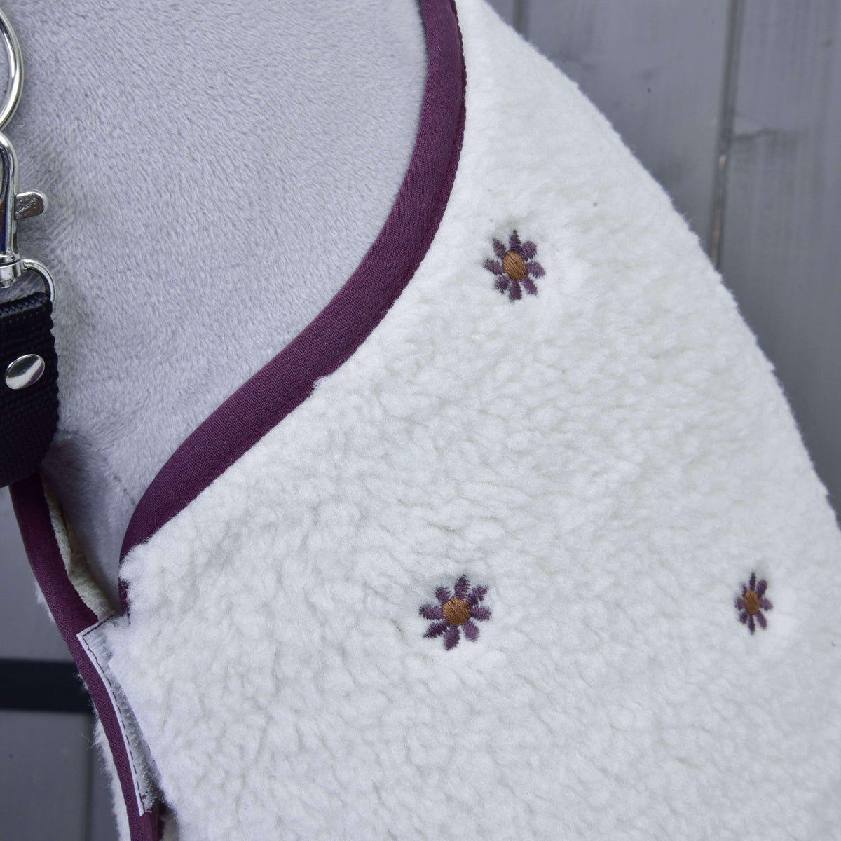 Detail Set Loret, bieleho fleecového psieho kabátika od Hobby Horse LarDen, s fialovo-žlto vyšitými kvetmi a fialovým lemom. Zobrazený na psovi s čiernym vodidlom pripomína deku pre Hobby Horse a leží na sivom drevenom povrchu.