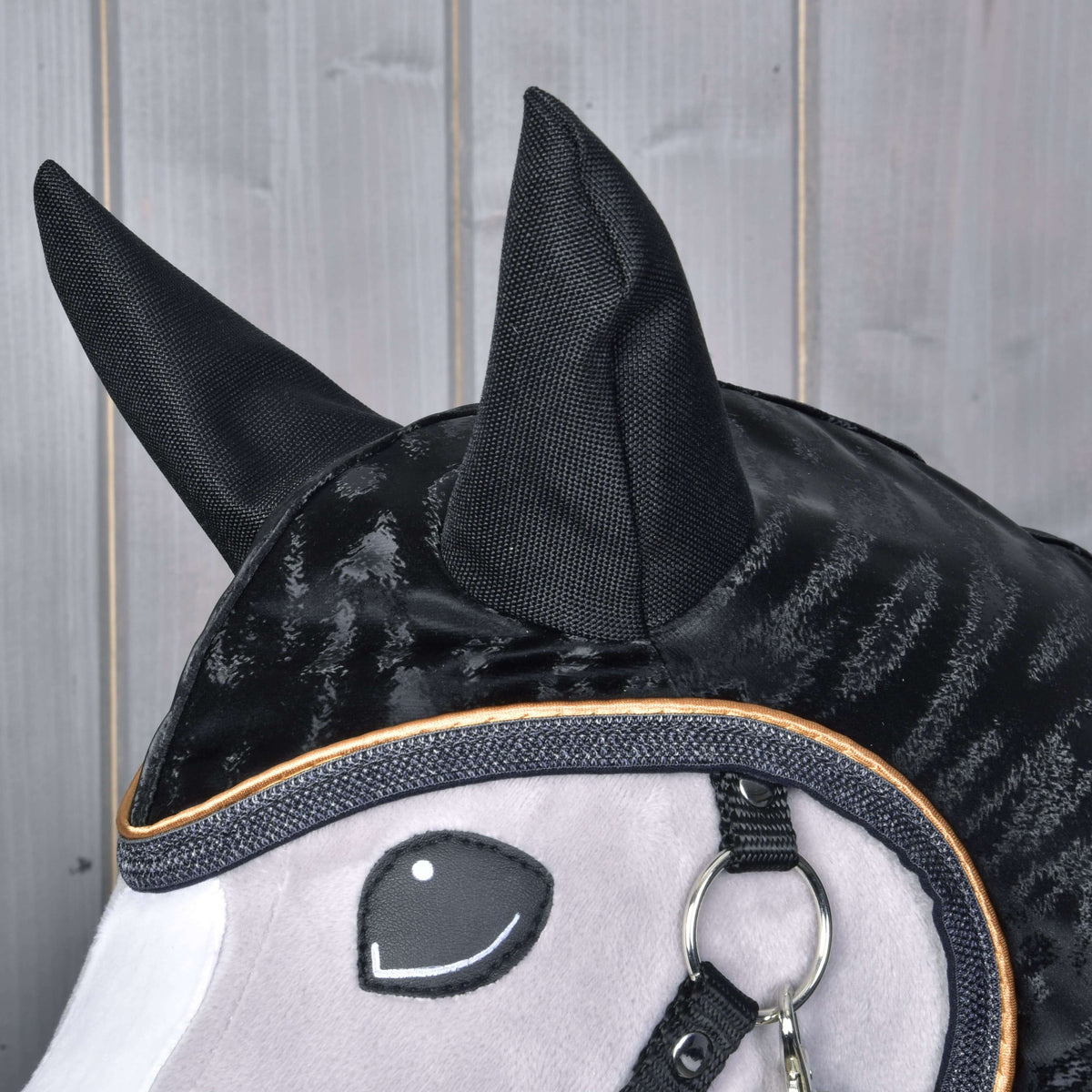 Detail Hobby Horse LarDen Pláštenka Luxury: čierno-sivá látková hlava Hobby Horse s veľkými čiernymi ušami, vyšitým usmievavým okom, lesklou čiernou hrivou a kovovým krúžkom pri papuli na pozadí drevených panelov.