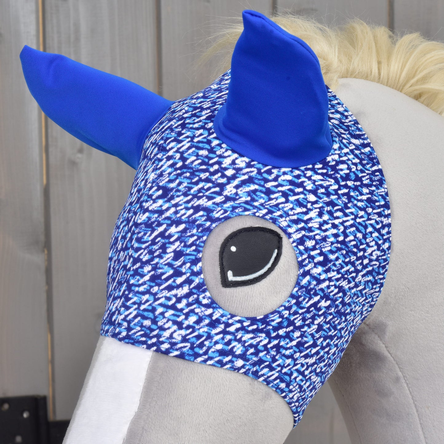Maska See od Hobby Horse LarDen, detail hlavy plyšového koňa s modrými ušami a modrobielym vzorom na drevenom pozadí.