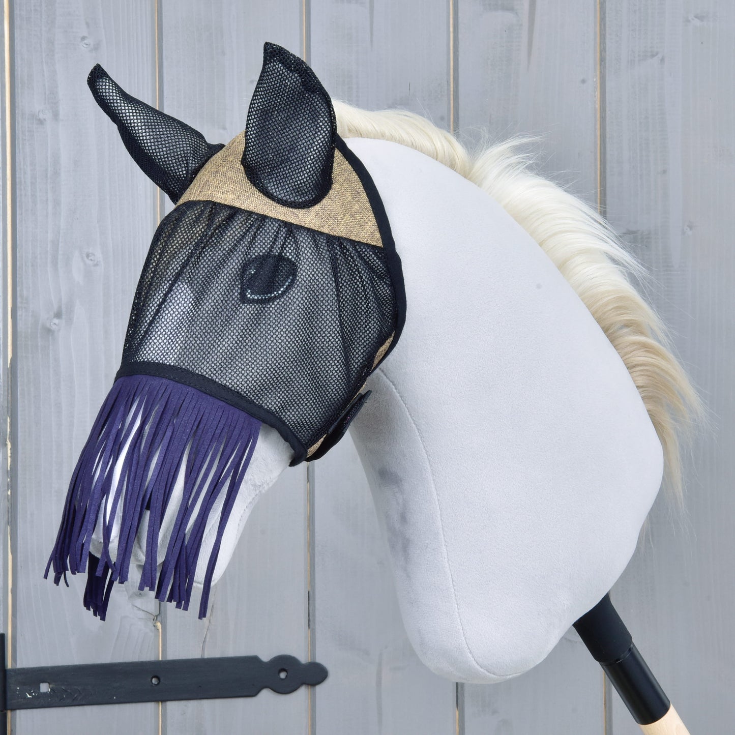 Maska proti hmyzu de Luxe Mona od Hobby Horse LarDen na bielom plyšovom Hobby Horse s čiernou tyčou a svetlo blond hrivou, so štýlovou strapcovou maskou proti hmyzu na pozadí sivej drevenej steny.