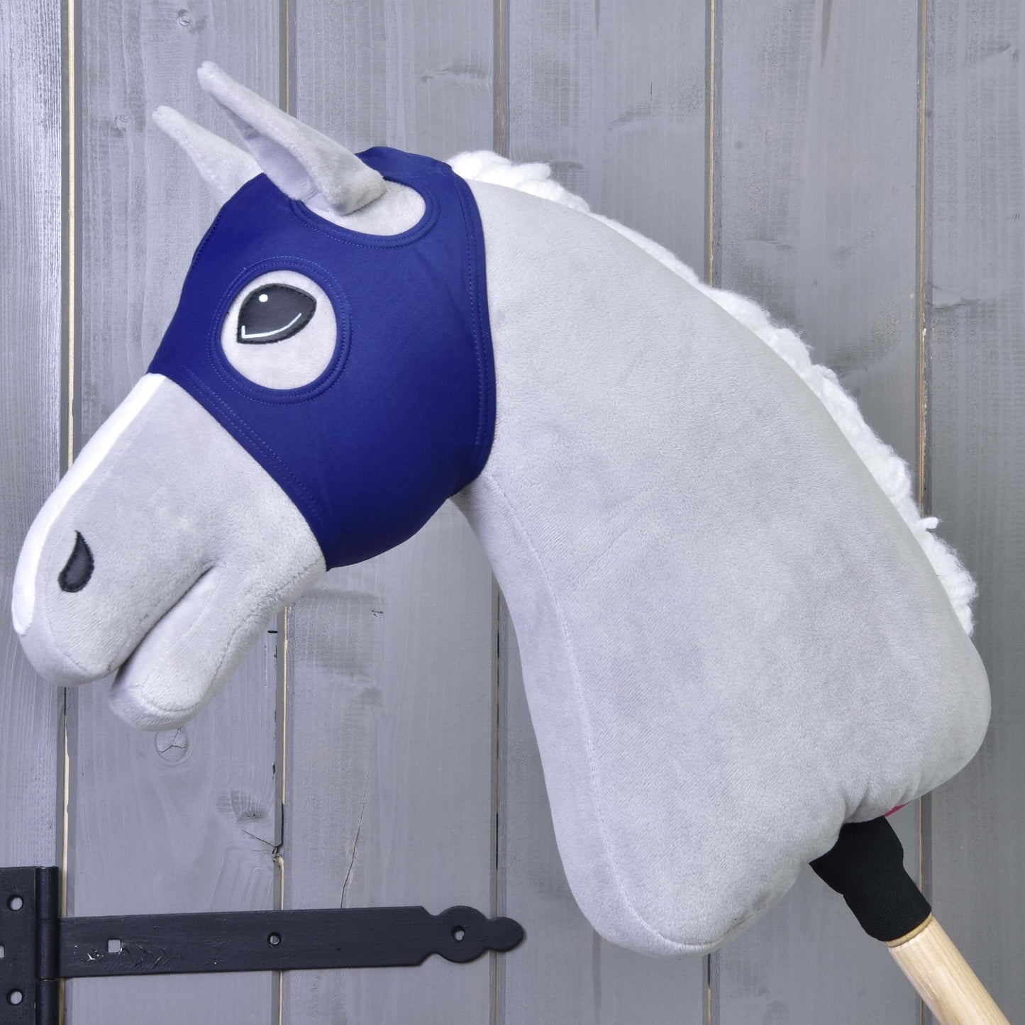 Sivý Hobby Horse LarDen s bielou hrivou a drevenou tyčou je zobrazený s maskou bez uší LarDen pred drevenou stenou s viditeľnými pántmi.