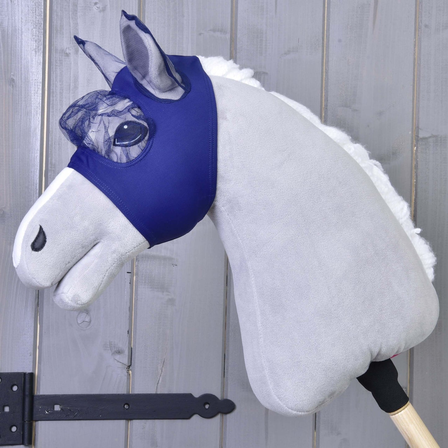 Maska proti hmyzu LarDen od Hobby Horse LarDen zobrazuje plyšového Hobby Horse so svetlosivým telom, bielou hrivou a prateľnou maskou proti hmyzu na drevenej tyči, na pozadí drevených panelov.