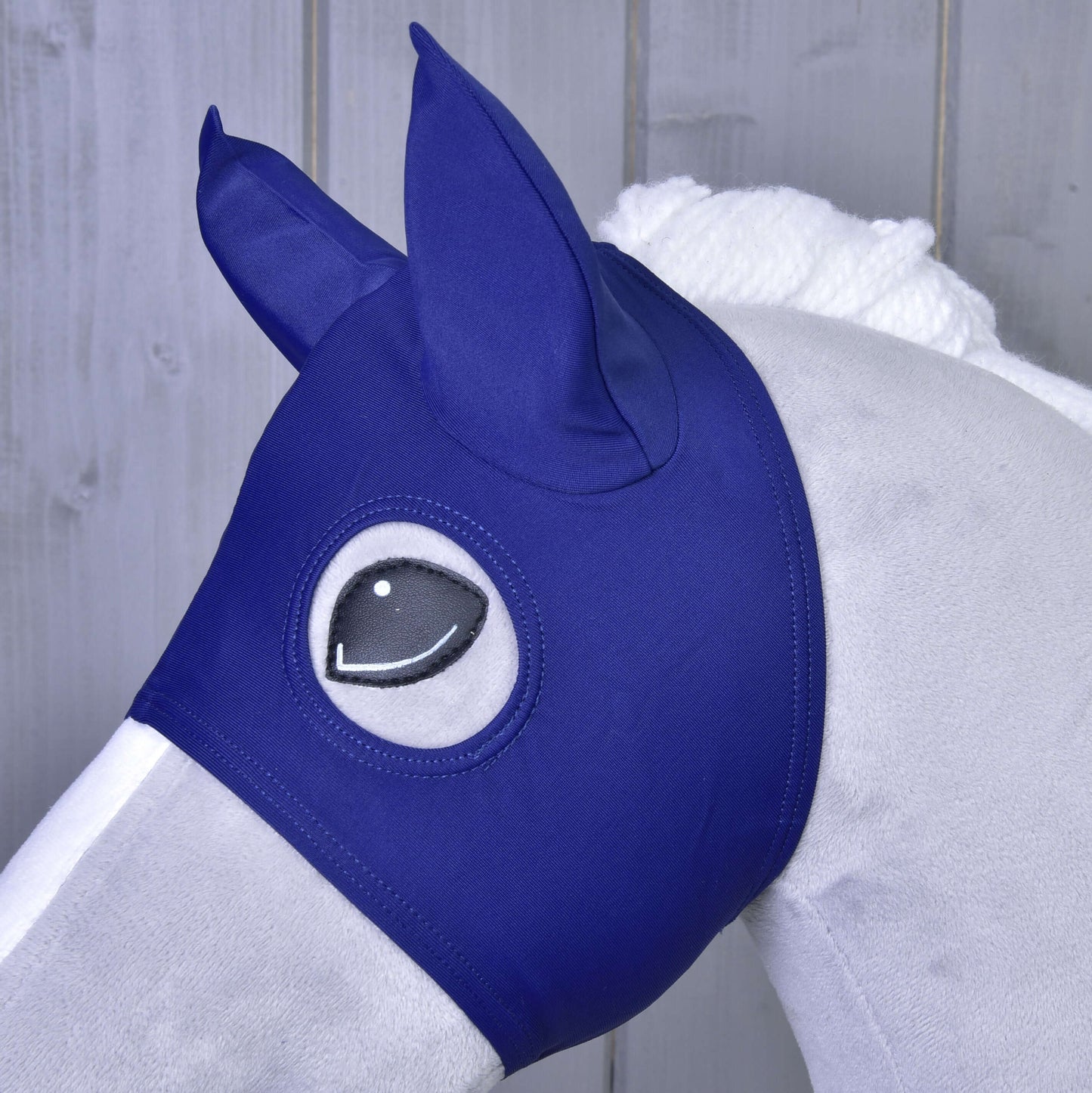 Detail sivého koňa s maskou Maska s ušami LarDen od Hobby Horse LarDen, s krytmi uší a otvormi pre oči, stojaceho pred dreveným pozadím.