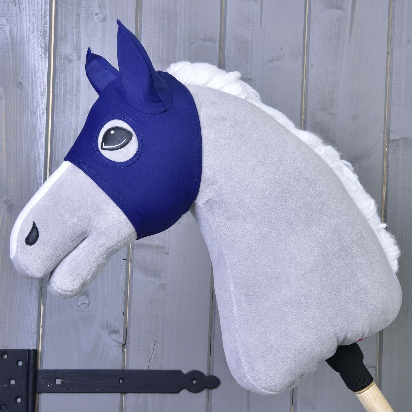 Maska s ušami LarDen od Hobby Horse LarDen na sivom plyšovom Hobby Horse s modrou látkovou uzdečkou a bielou hrivou na drevenej tyči – ideálna pre nápaditú hru PES, zobrazená pred sivou drevenou stenou.
