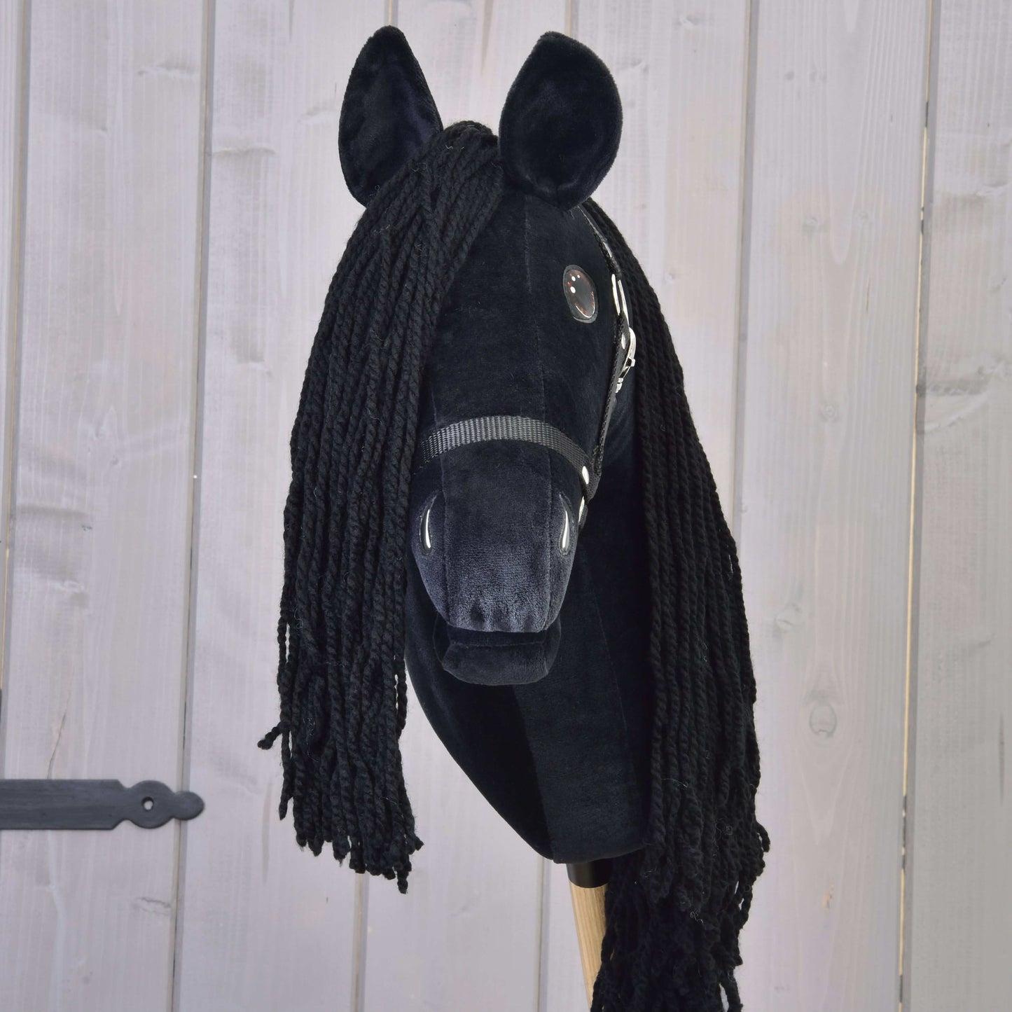 Hobby Horse Maximus s čiernou ohlávkou od Hobby Horse LarDen, ručne vyrábaný frízsky Hobby Horse s plyšovou hlavou, priadzovou hrivou, gombíkovými očami, čiernou ohlávkou a drevenou tyčou, zobrazený na svetlom drevenom pozadí.