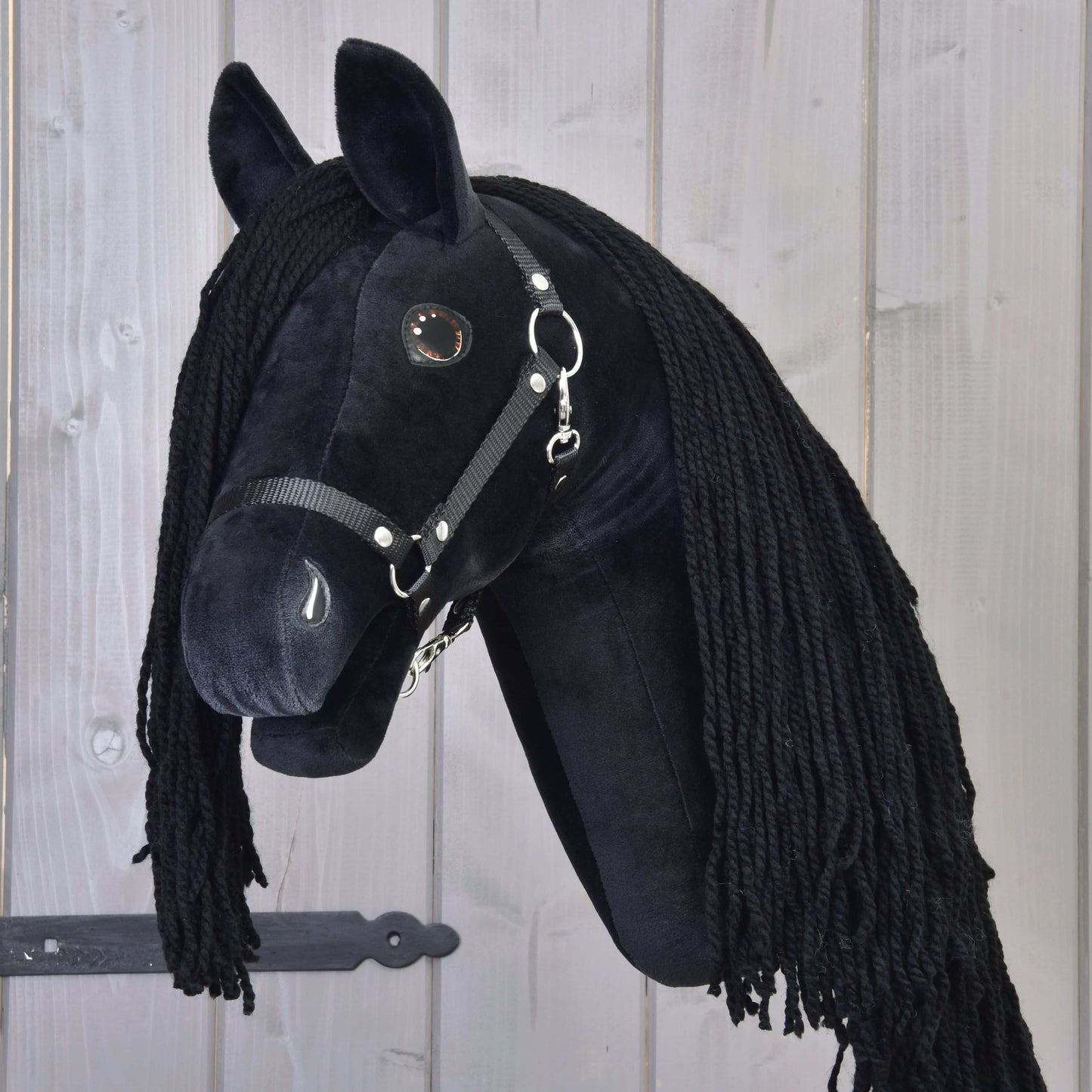 Hobby Horse Maximus s čiernou ohlávkou od Hobby Horse LarDen, plyšový čierny frízsky Hobby Horse s dlhou čiernou hrivou, na drevenej tyči, zobrazený pred svetlým dreveným obložením.