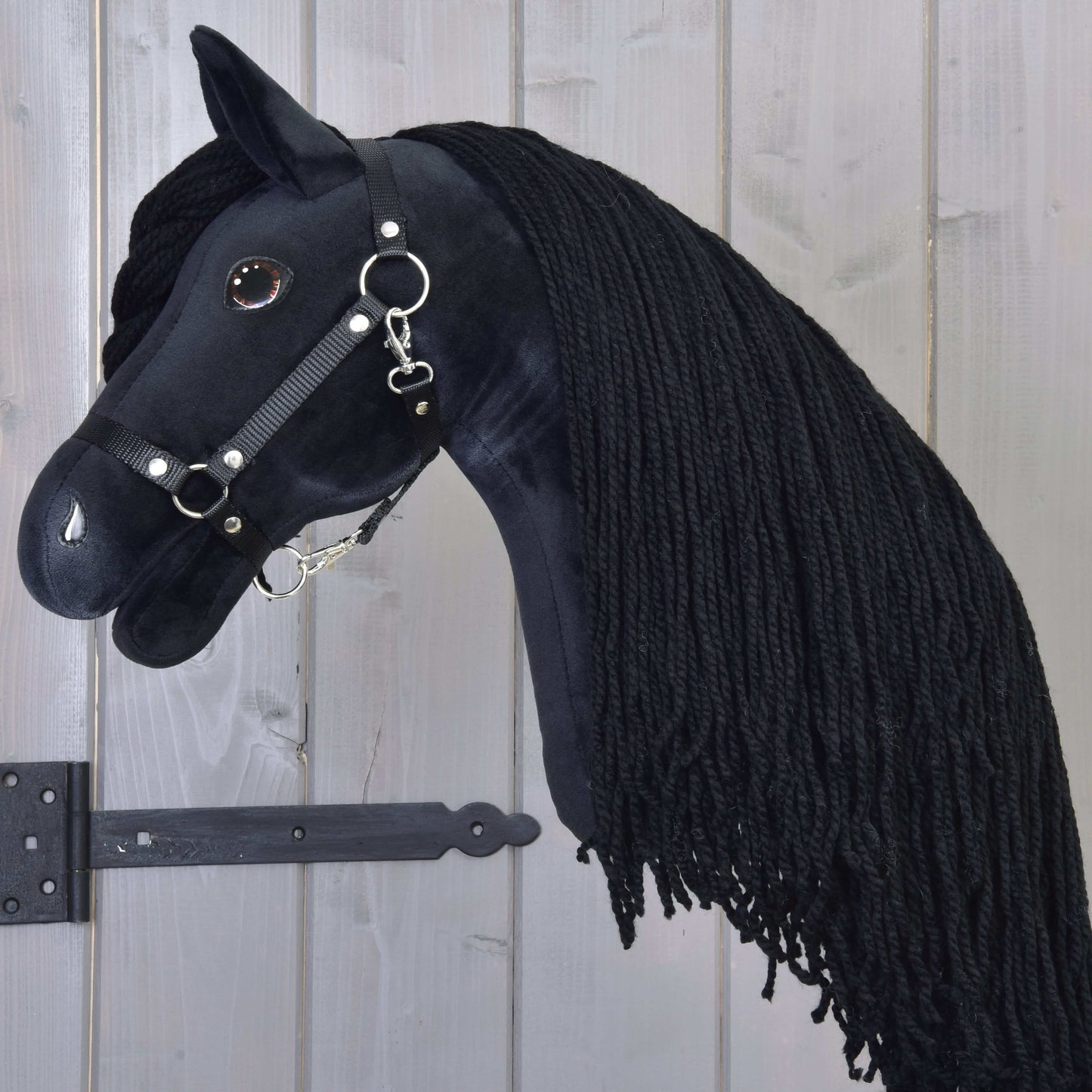 Hobby Horse Maximus s čiernou ohlávkou od Hobby Horse LarDen s plyšovým čiernym frízskym vzhľadom, dlhou zapletenou hrivou, uzdečkou s kovovými krúžkami a lesklým gombíkovým okom, zavesený na drevenej stene.