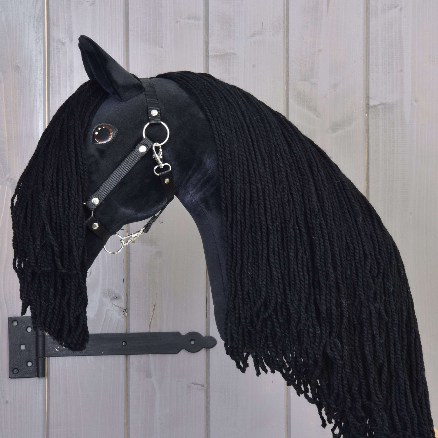 Hobby Horse Maximus s čiernou ohlávkou od Hobby Horse LarDen, ručne vyrábaná hračka čierneho žrebca s dlhou priadzovou hrivou, lesklou čiernou uzdečkou a gombíkovým okom, upevnená na dreve s čiernym kovovým držiakom.