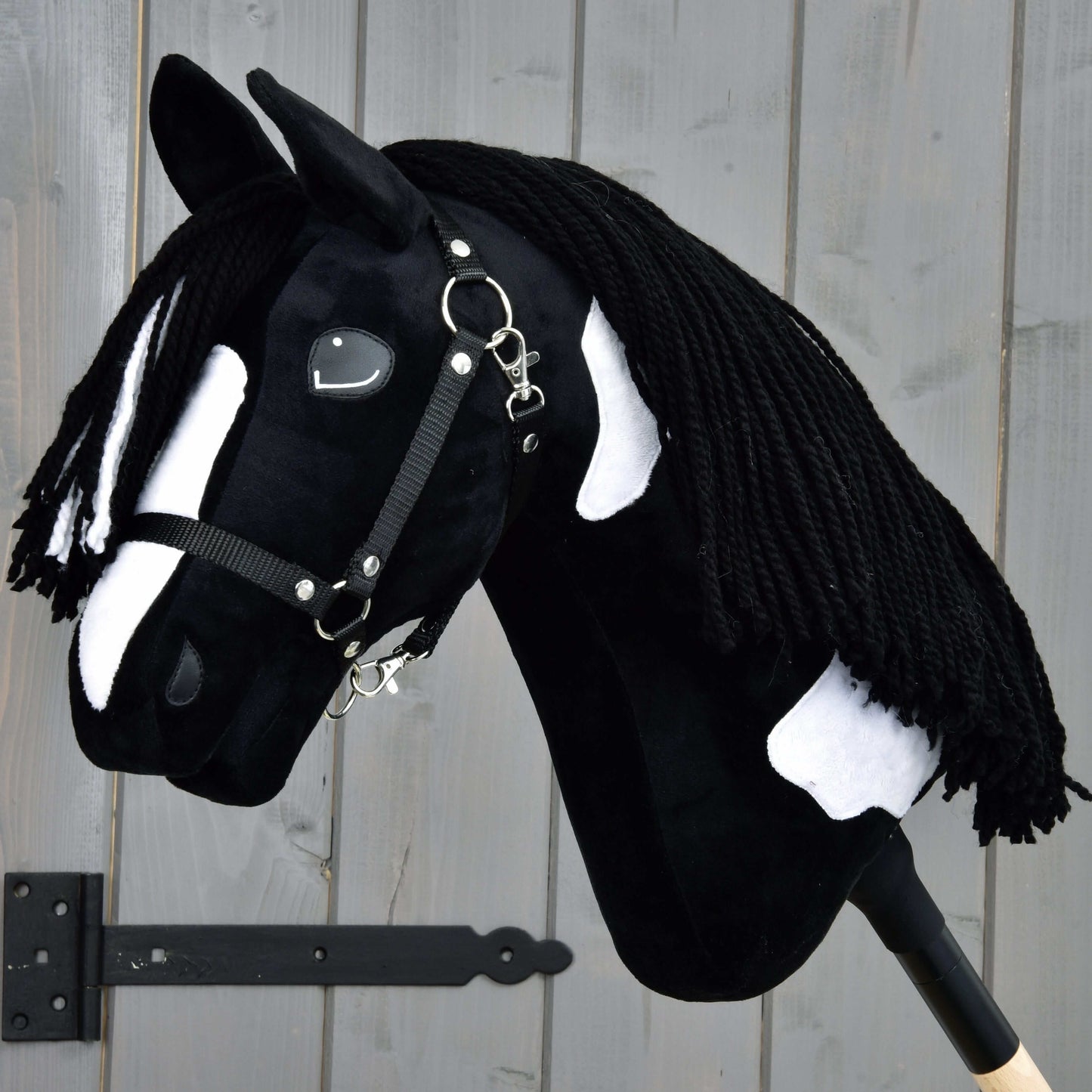 Hobby Horse Merlin s čiernou ohlávkou od Hobby Horse LarDen, inšpirovaný žrebcom plemena Barock Pinto, má čiernobiele škvrny a zapletenú priadzovú hrivu, upevnený pred sivou drevenou stenou.