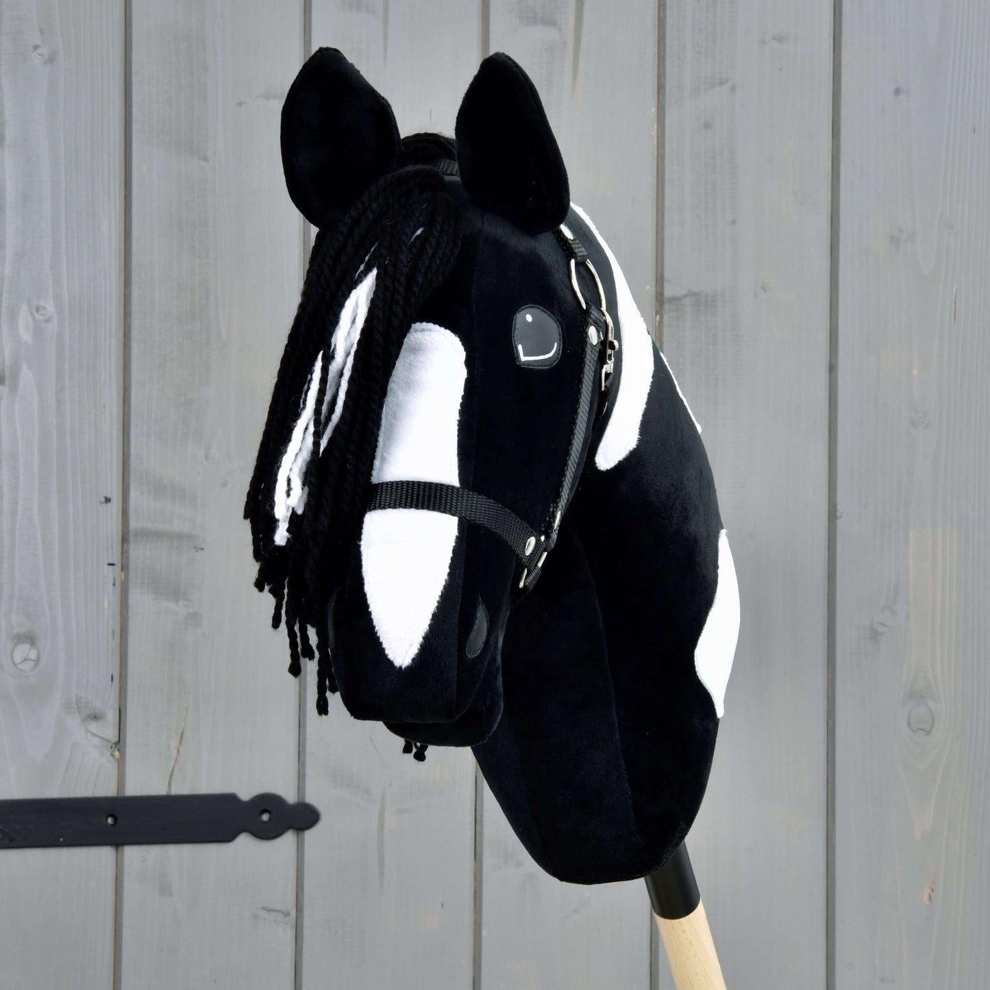 Hobby Horse Merlin s čiernou ohlávkou od Hobby Horse LarDen je čiernobielo škvrnitý Hobby Horse v štýle žrebca Barock Pinto, s priadzovou hrivou a uzdečkou, upevnený na drevenej tyči pred sivým plotom.