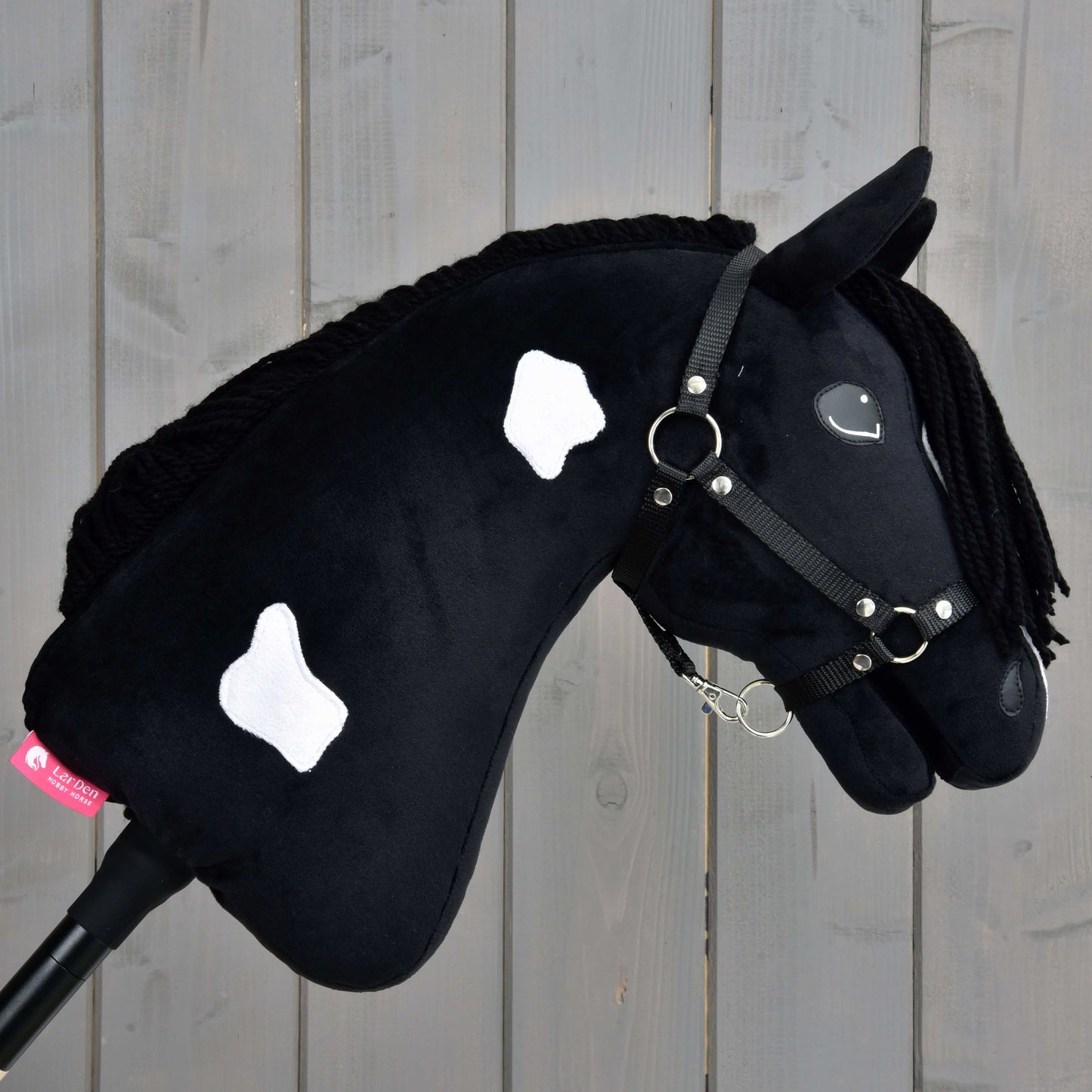 Hobby Horse Merlin s čiernou ohlávkou od Hobby Horse LarDen má čierne telo inšpirované Barock Pinto s bielymi škvrnami, zapletenú hrivu a čiernu ohlávku, na pozadí drevených dosiek.