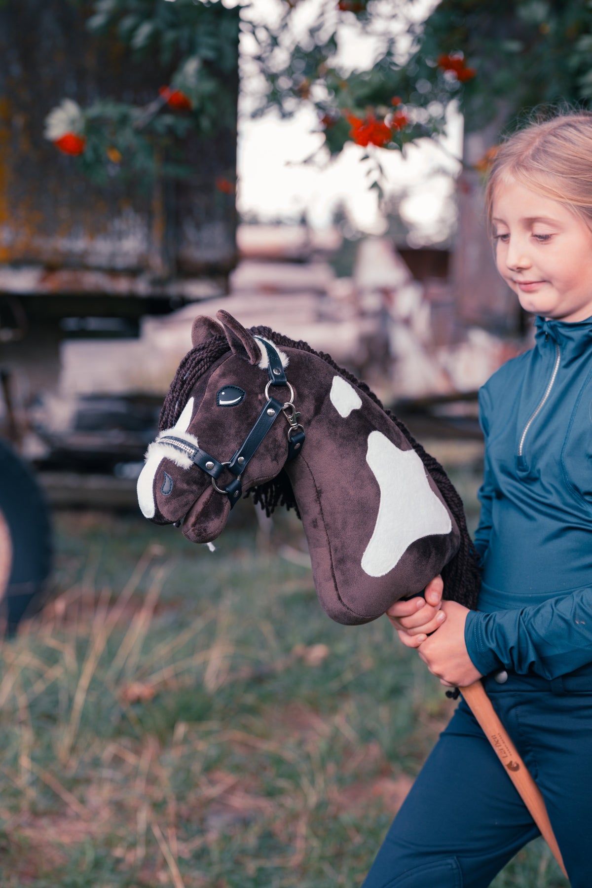 Mladý účastník v tmavomodrom jazdeckom odeve drží Hobby Horse Monte od Hobby Horse LarDen na lúke pred starým dreveným vozom a jarabinou s červenými bobuľami, s hnedo-bielou srsťou inšpirovanou plemenom American Paint Horse, dlhou tmavou priadzovou hrivou a ohlávkou Espresso s kožušinovým lemom