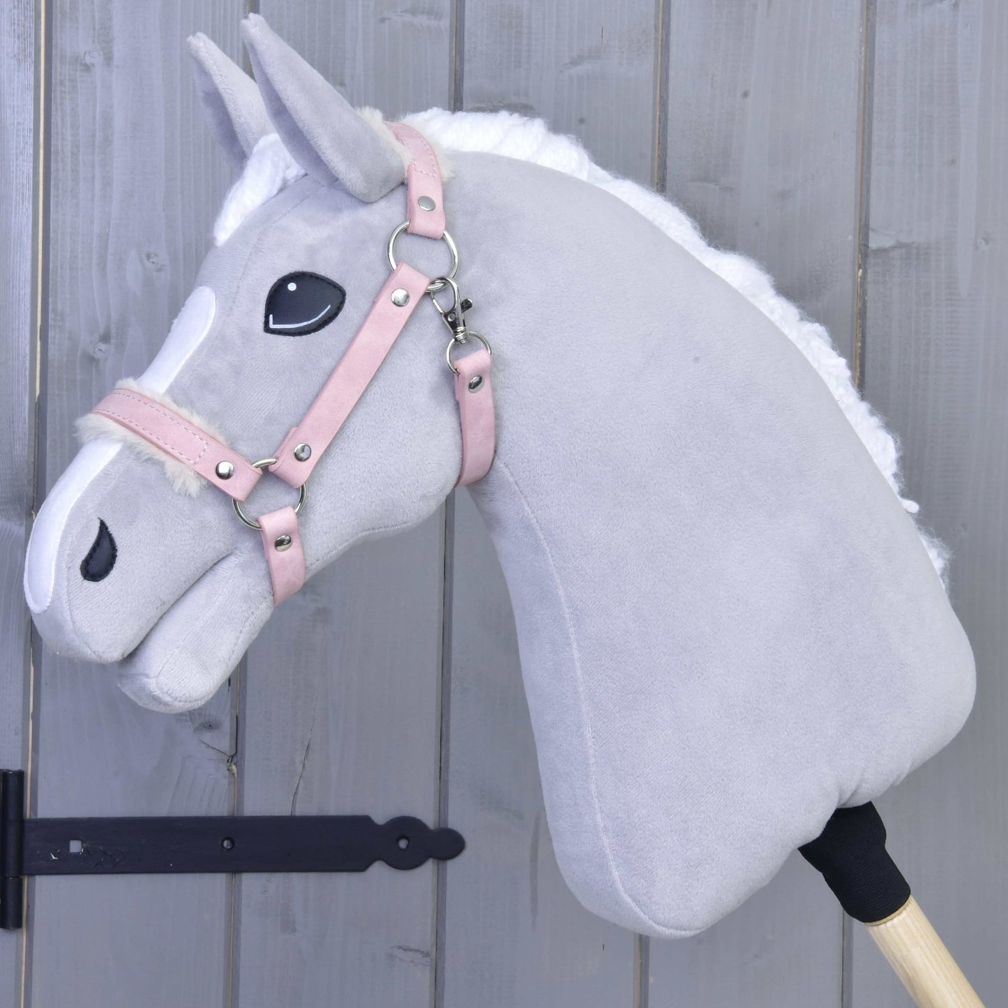 Hobby Horse LarDen Ohlávka s barančekom z koženky Mullbery na svetlosivom plyšovom Hobby Horse s bielou hrivou, ružovou uzdečkou z koženky a vyšívanými detailmi, nasadenom na drevenej tyči pred sivou drevenou stenou.