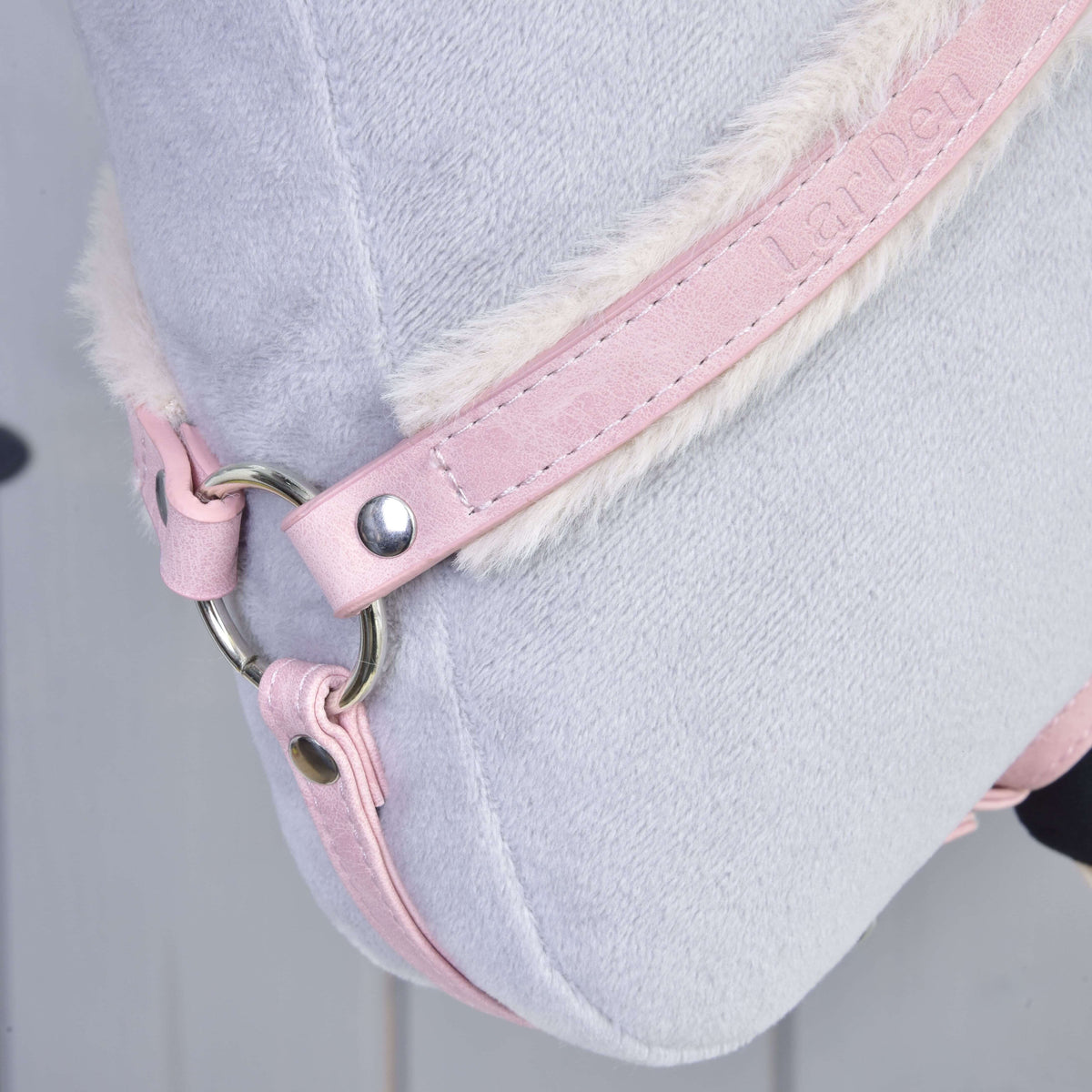 Detail Poprsníka s barančekom Mullbery od Hobby Horse LarDen so svetlosivým plyšom, ružovým koženkovým remienkom s razeným textom, bielym lemom z umelej kožušiny a kovovým krúžkom. Pozadie je rozmazané.