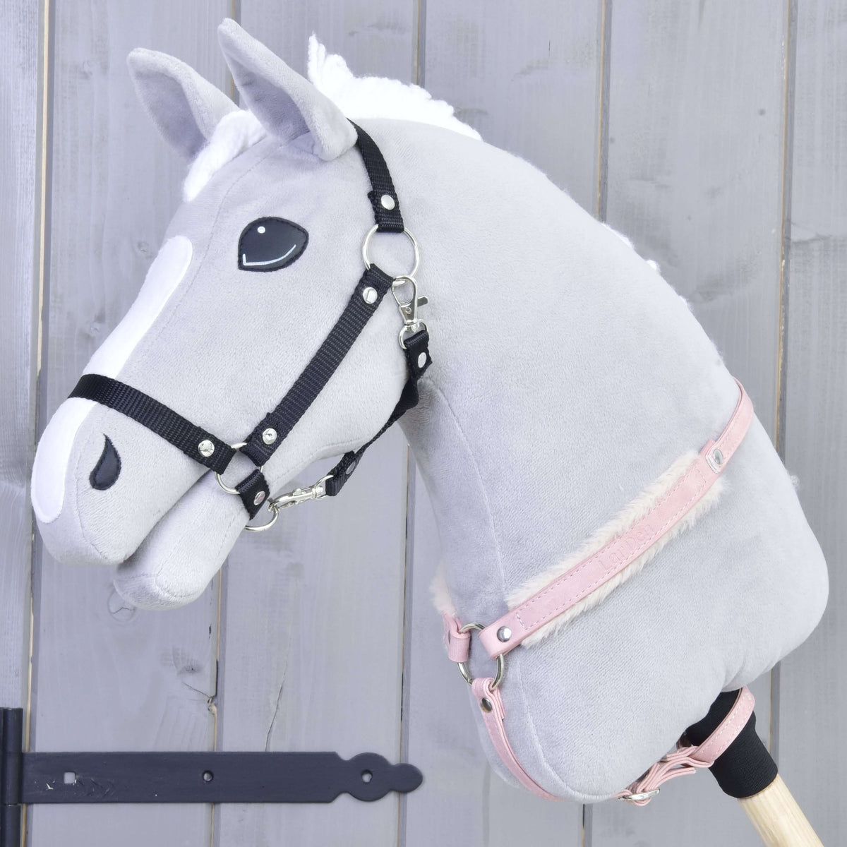 Poprsník s barančekom Mullbery od Hobby Horse LarDen na sivom plyšovom Hobby Horse s bielou hrivou, čiernou uzdečkou, ružovou ohlávkou a mäkkým poprsníkom s kožušinovou podšívkou na drevenej tyči, na svetlosivom drevenom pozadí.