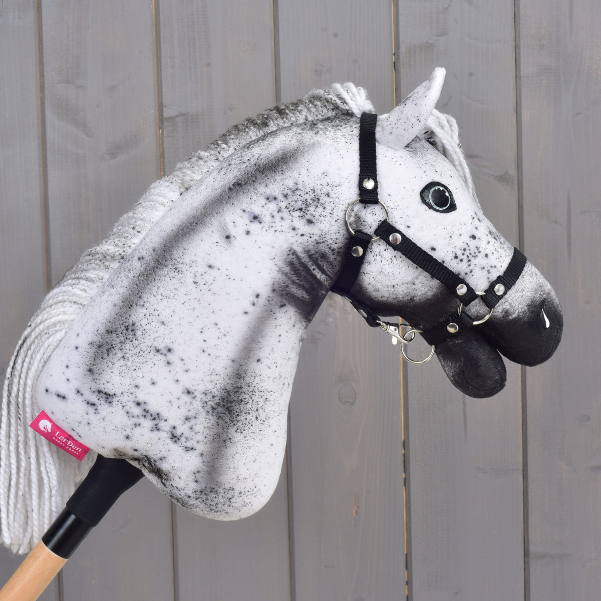 Hobby Horse Naivy s čiernou ohlávkou - veľkosť S od Hobby Horse LarDen, s výrazným bielo-sivým škvrnitým vzhľadom, hrdo stojí nasadený na drevenej tyči pred sivou drevenou stenou.