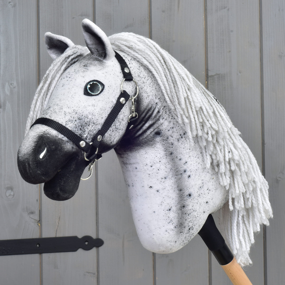 Hobby Horse LarDen "Hobby Horse Naivy with Black Halter" (veľkosť S), so sivo-bielym appaloosa vzhľadom, čiernou ohlávkou a priadzovou hrivou, nasadený na drevenej tyči a umiestnený pred sivou drevenou stenou.