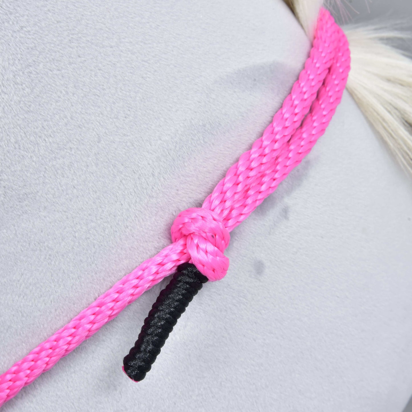 Detail Nákrčníka Pink Festive od Hobby Horse LarDen – výrazne ružové ručne vyrobené cordeo lano s prameňom s čiernym zakončením, uviazané do uzla okolo svetlosivého povrchu. Ideálne na jazdenie bez uzdečky; pozadie je hladké s jemnými stopami.