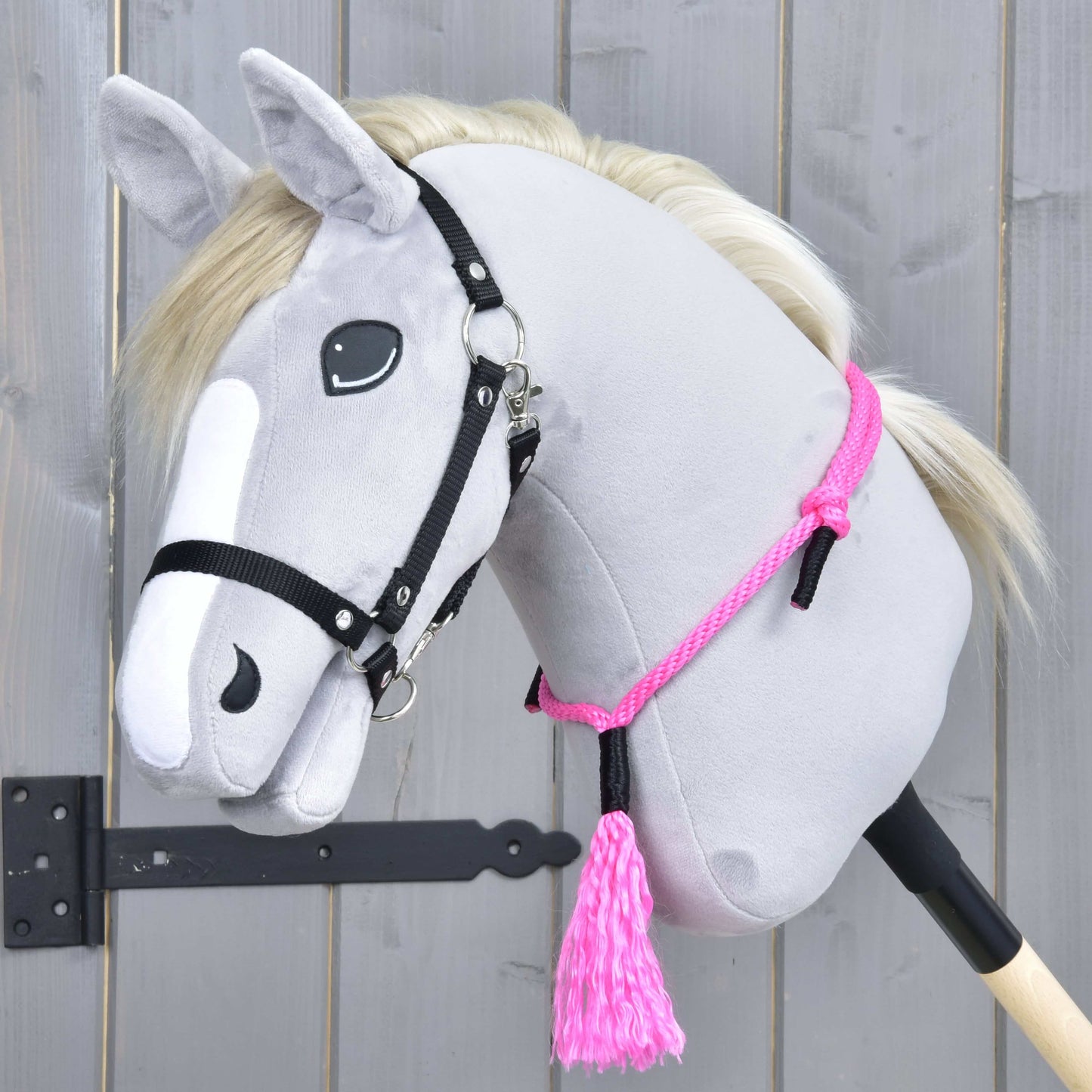 Hobby Horse LarDen „Nákrčník Pink Festive“ na plyšovom sivom Hobby Horse s bielou hrivou, čiernou uzdečkou, ručne vyrobeným cordeom, ružovým lanovým nákrčníkom a ružovým strapcom, na drevenom pozadí.