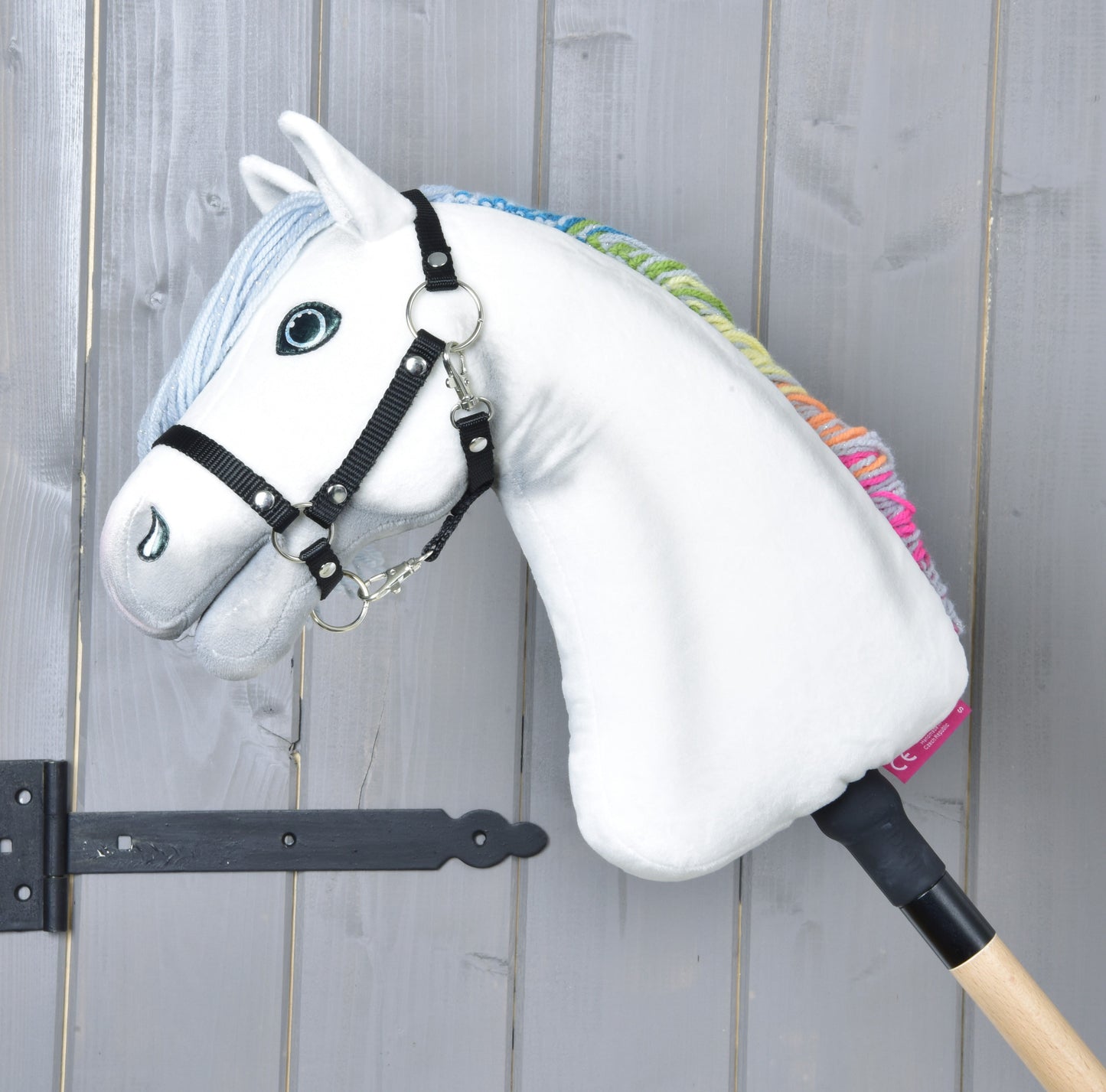 Hobby Horse Rainbow New od Hobby Horse LarDen, biely hračkársky kôň s čiernou uzdečkou a dúhovou hrivou na drevenej tyči, opretý o sivú drevenú stenu.