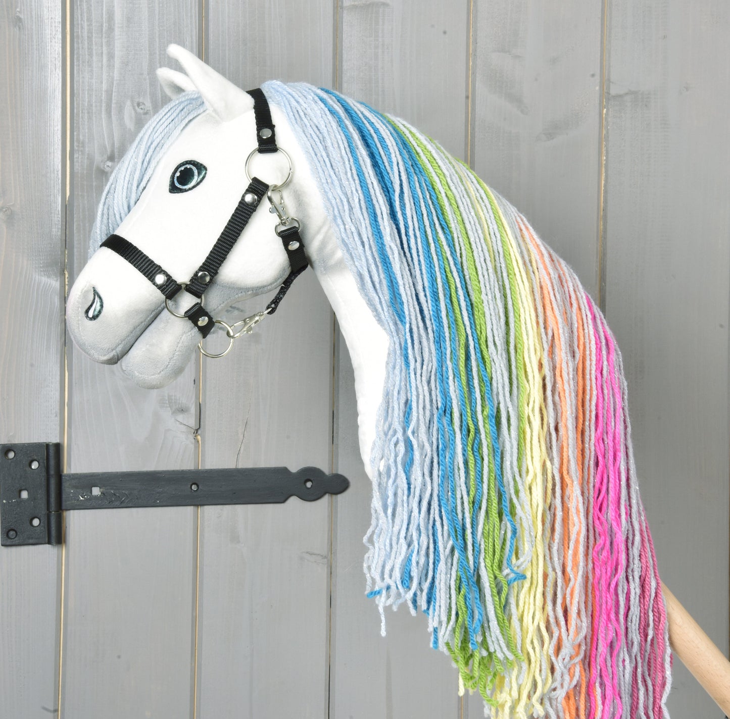Hobby Horse LarDen „Hobby Horse Rainbow New“ má bielu tvár, čiernu uzdečku a dlhú dúhovo sfarbenú hrivu z priadze, stojí pred sivými drevenými dverami.