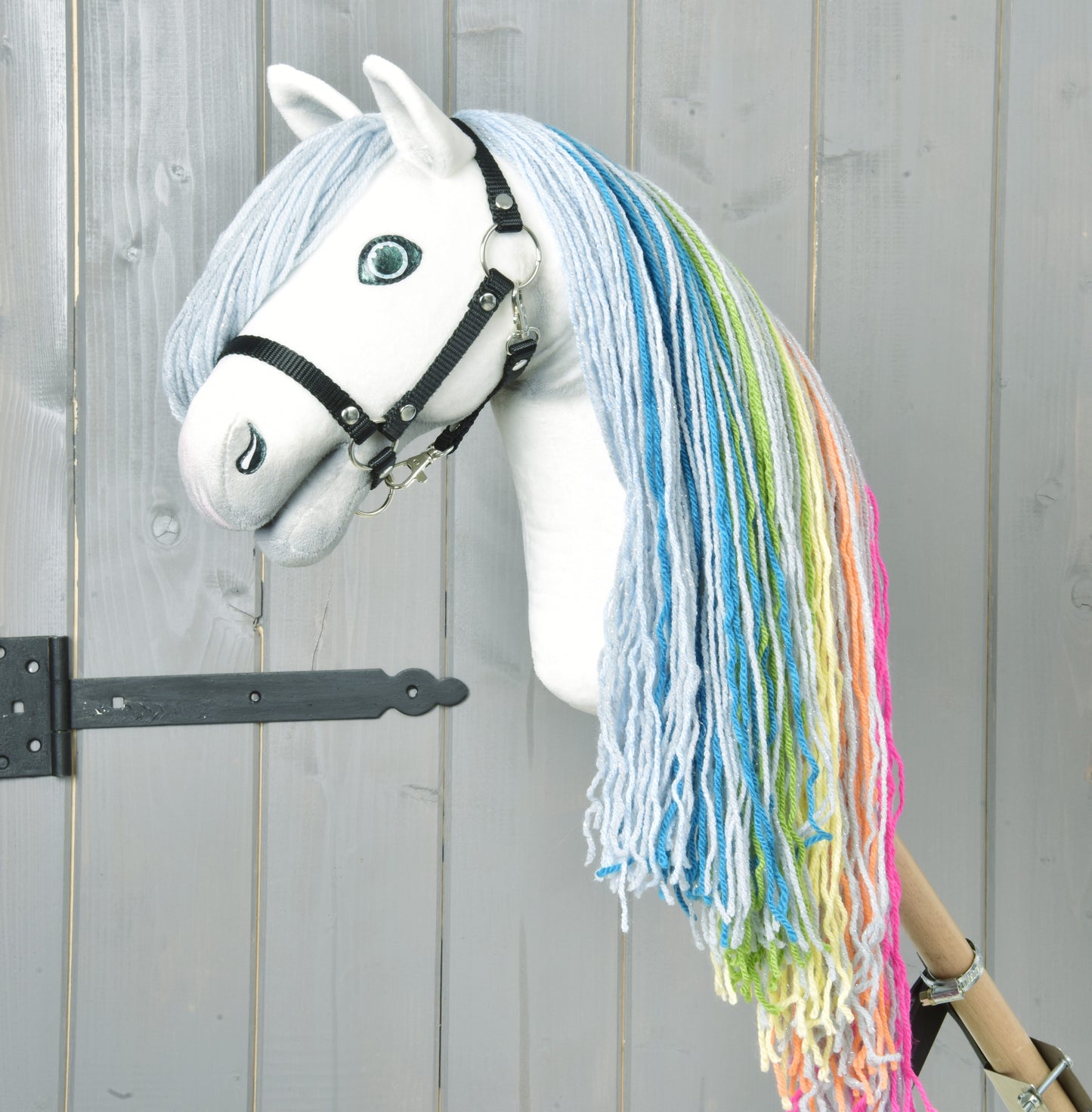 Hobby Horse Rainbow New od Hobby Horse LarDen má ručne vyrobenú bielu tvár, modré oči, čiernu uzdečku a dúhovú hrivu z modrej, zelenej, žltej a ružovej priadze na svetlosivom drevenom pozadí pre rozprávkový dojem.