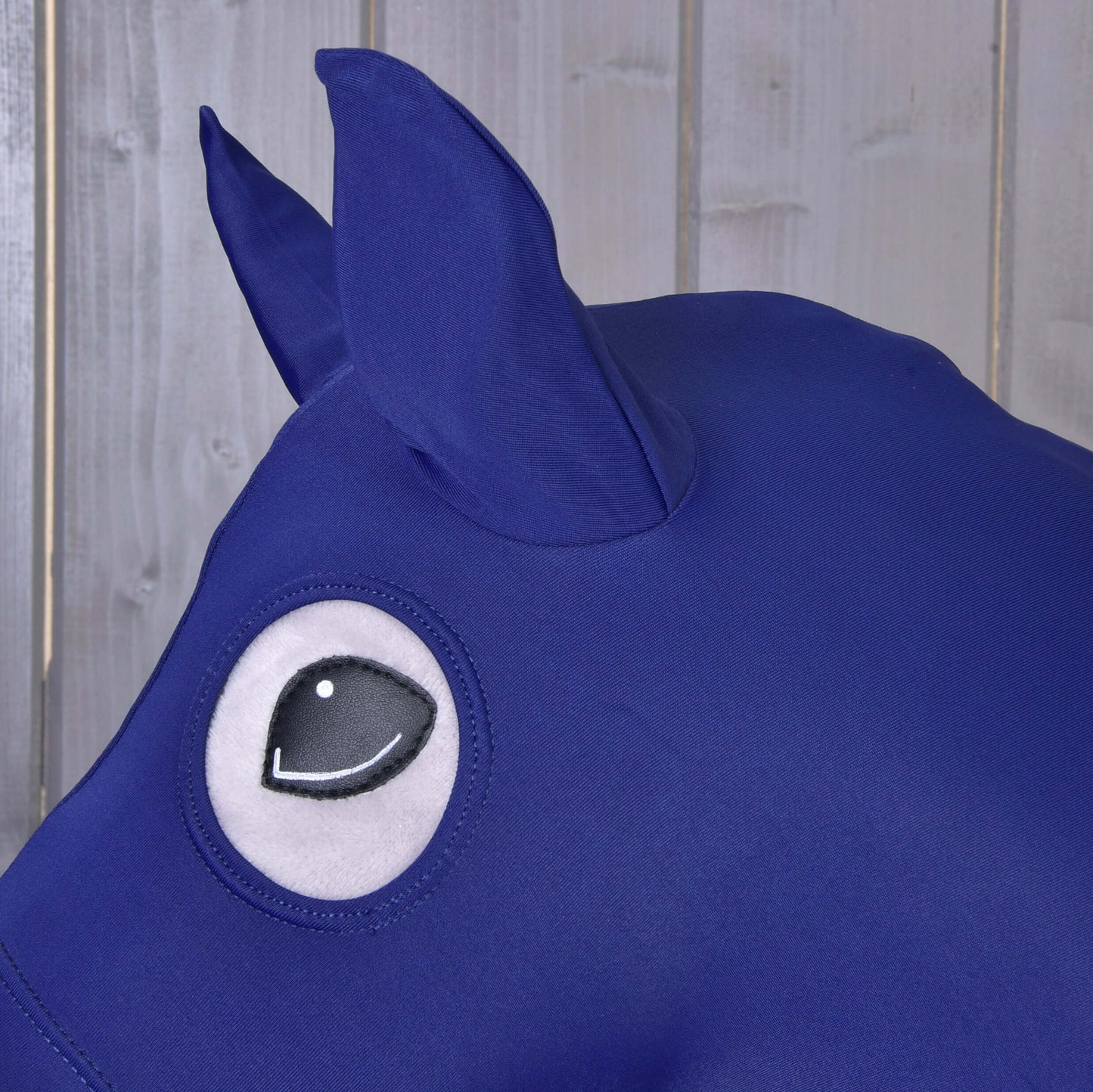 Detail modrého ochranného obleku Hobby Horse LarDen z Lycry, navrhnutého pre Hobby Horse, s veľkým okom v kreslenom štýle a špicatým uchom, zobrazeného na pozadí svetlých drevených panelov.