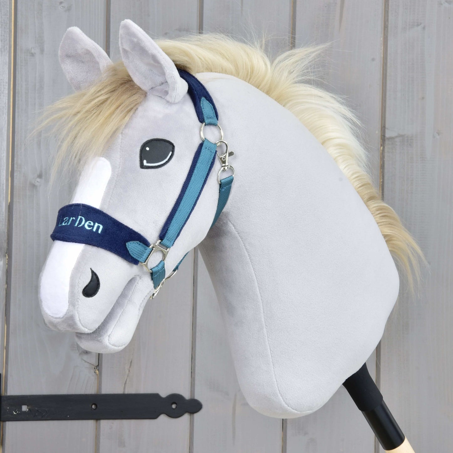 Hobby Horse LarDen s ohlávkou s barančekom LarDen Blue, so svetlosivou plyšovou hlavou, bielou hrivou a modrou kožušinovou ohlávkou, upevnený na tyči na drevenom pozadí.