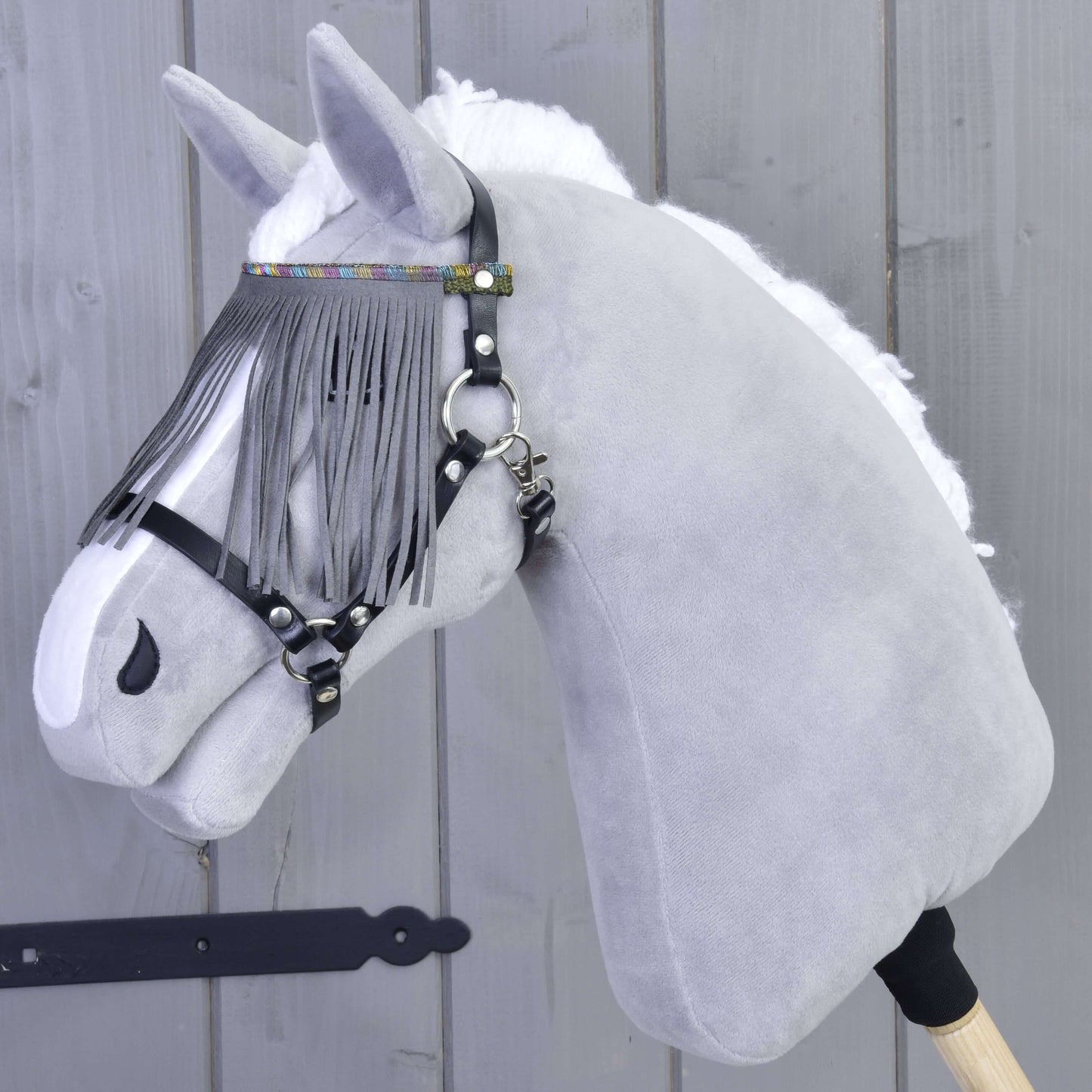 Plyšová sivá hlava Hobby Horse s bielou hrivou, s ohlávkou od Hobby Horse LarDen v čiernej farbe so sivými strapcami a sivým nánosníkom, na drevenej tyči, pred drevenou stenou.