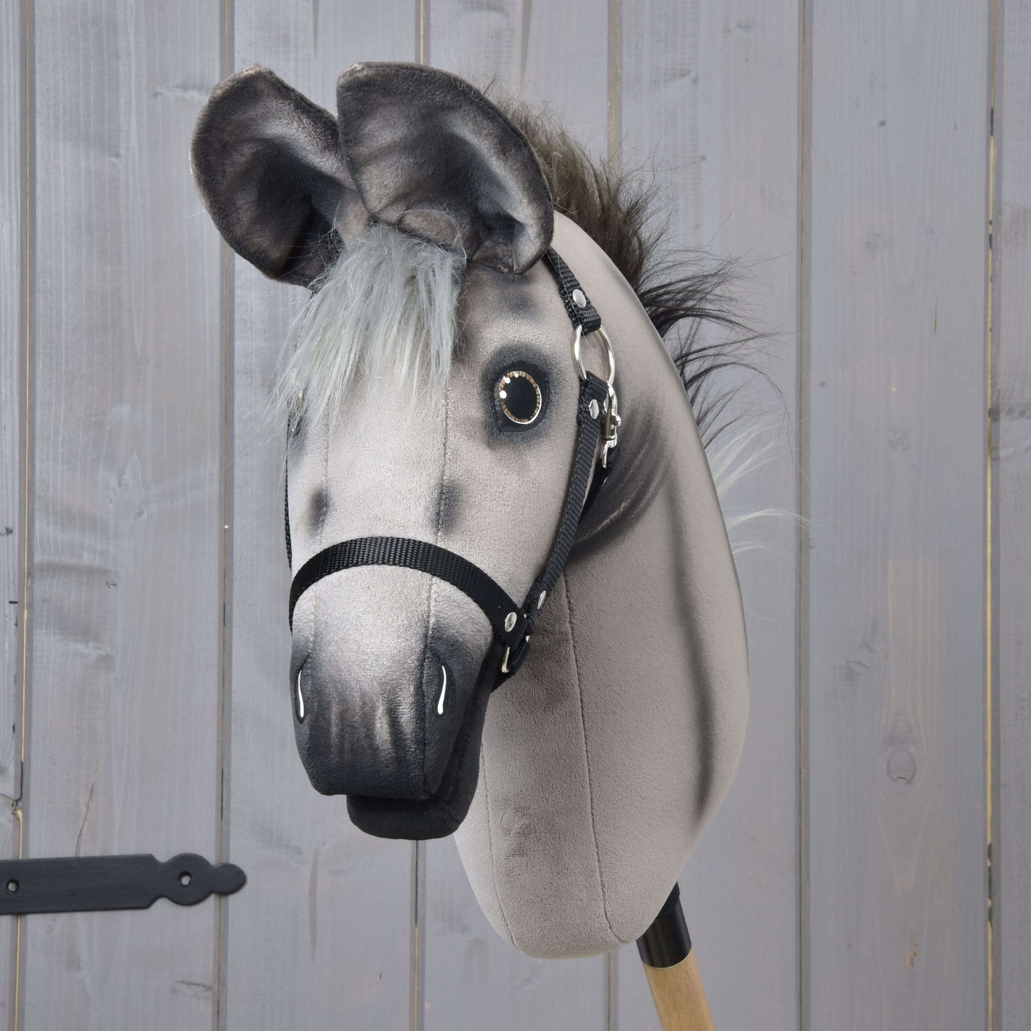 Hobby Horse osol Olin s čiernou ohlávkou od Hobby Horse LarDen, sivá oslia hračka na drevenej tyči s maľovanými detailmi a čiernym postrojom, pred svetlosivým dreveným obkladom.