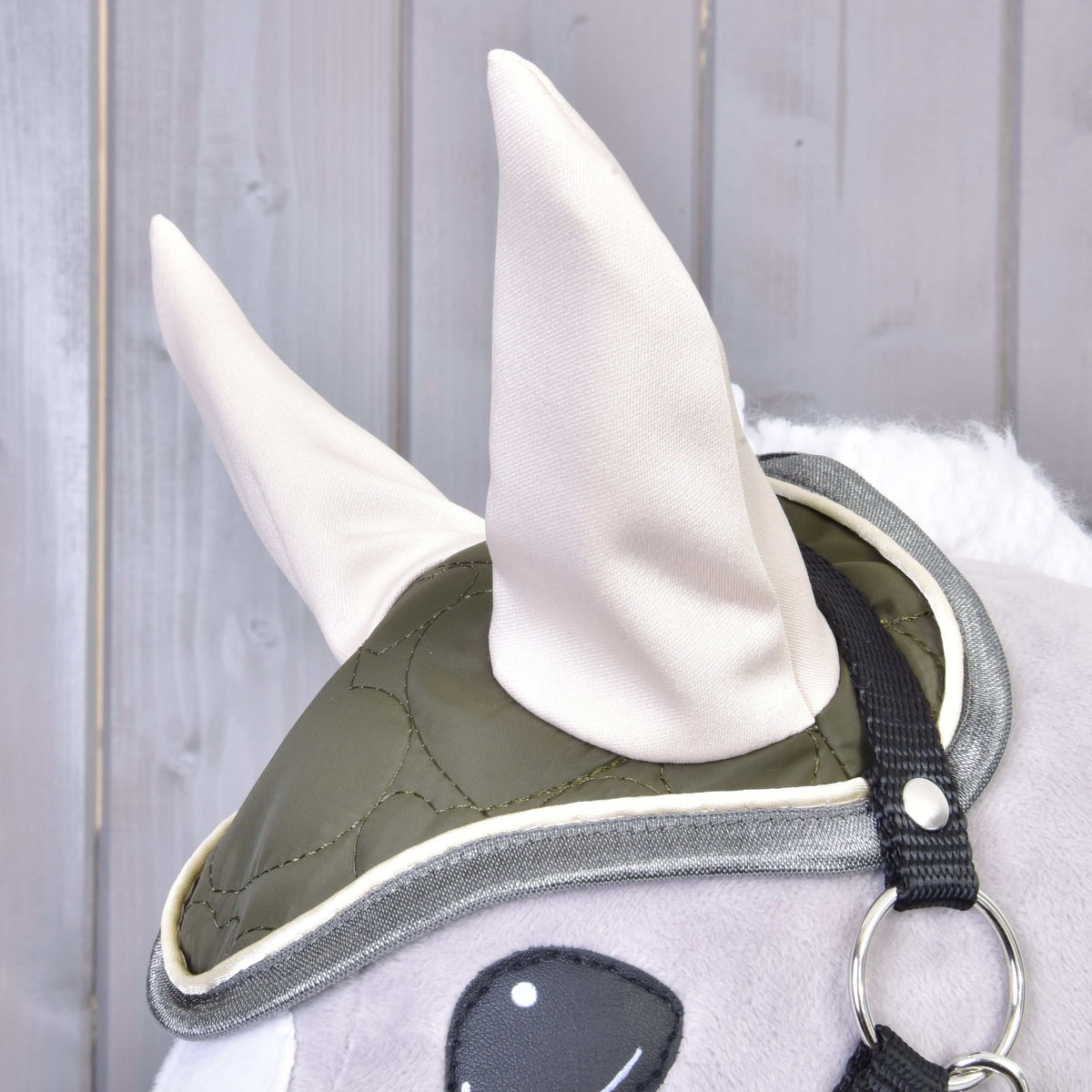 Detail plyšového koňa so Set Olivetti od Hobby Horse LarDen: zelená prešívaná čabraka s dvoma veľkými svetlými ušami, čiernym popruhom a kovovým krúžkom – jedinečné konské doplnky na drevenom pozadí.