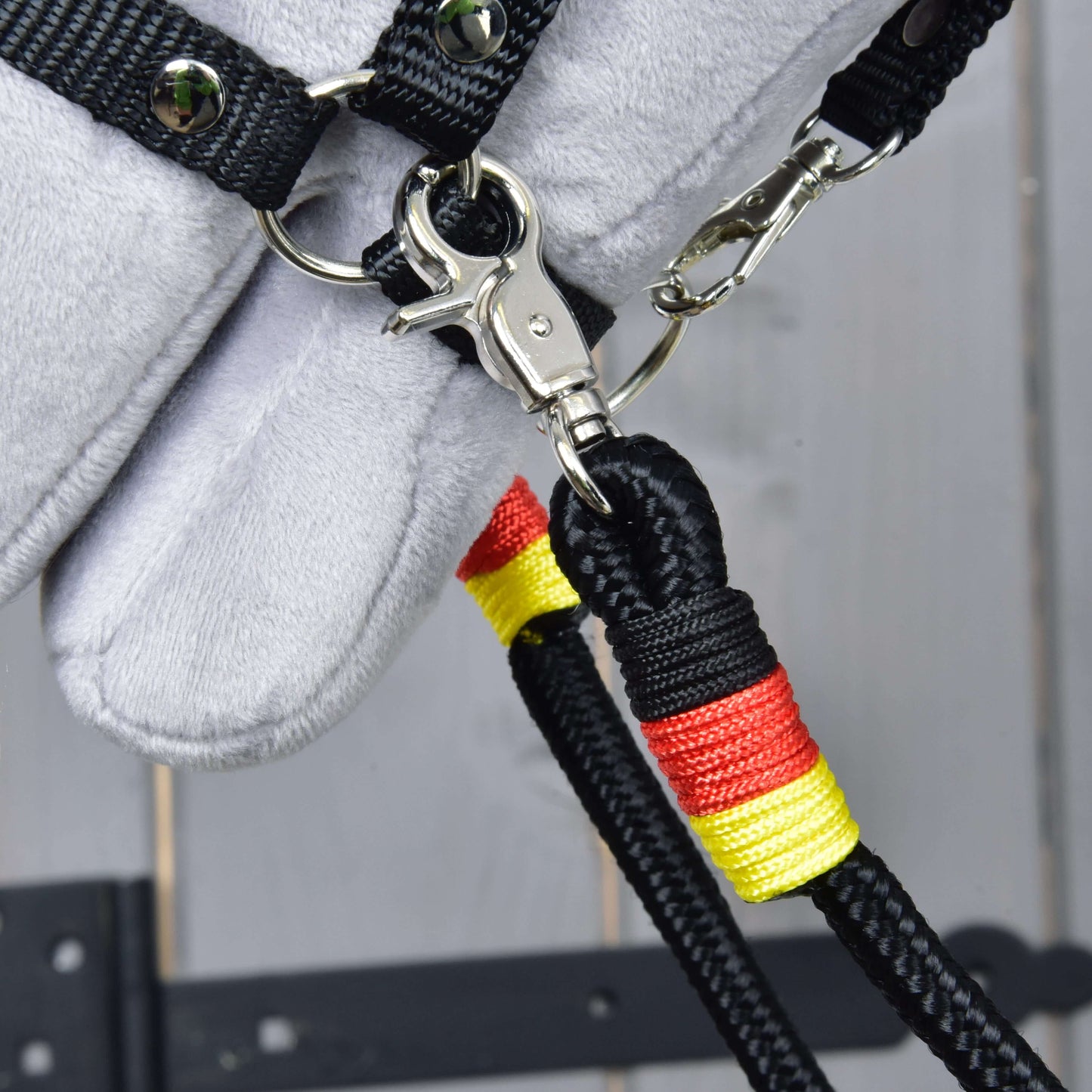 Detail oprát Opraty Emblem od Hobby Horse LarDen – čierne opraty z polypropylénového lana s červenou a žltou omotávkou, pripnuté ku kovovému krúžku na čiernom remienku, pripevnené k sivej plyšovej konskej hlave na drevenom pozadí.