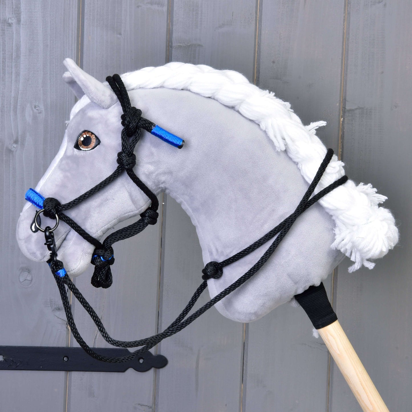 Parelka Azure od Hobby Horse LarDen na plyšovom sivom Hobby Horse s bielou priadzovou hrivou, čiernou uzdečkou a výraznými azúrovými akcentmi, nasadenom na drevenej tyči pred sivou drevenou stenou.