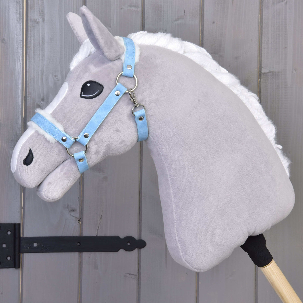 Sivý plyšový koník na tyči s bielou hrivou má na sebe Hobby Horse LarDen Ohlávka s barančekom z koženky Pastell Blue a je opretý o drevenú panelovú stenu s čiernymi kovovými pántmi.