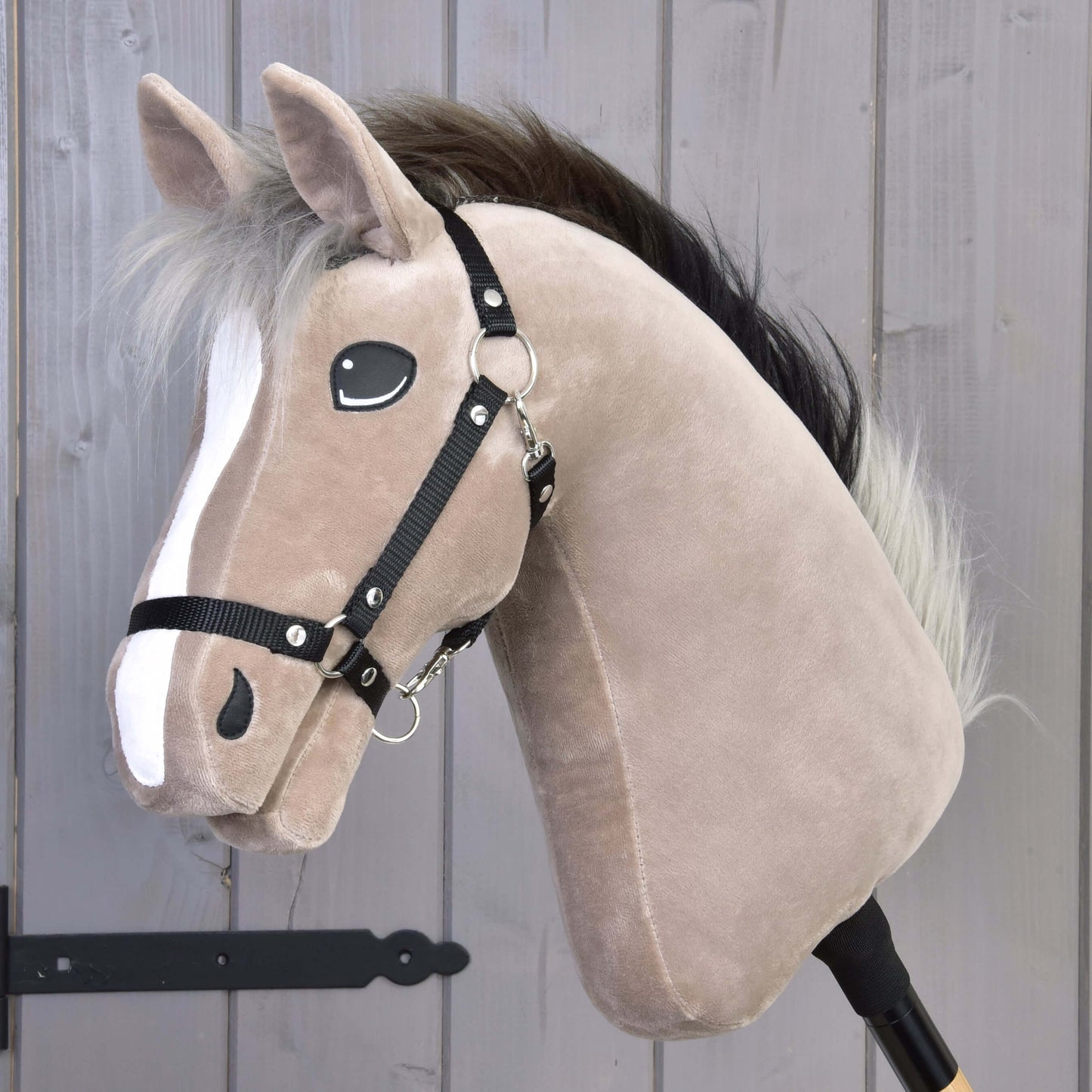 Hobby Horse Piccolino s čiernou ohlávkou od Hobby Horse LarDen, svetlohnedý plyšový športový kôň plemena holštajn s bielym pruhom na hlave, čiernou ohlávkou, huňatou hrivou a na drevenej tyči, pred sivou drevenou stenou.