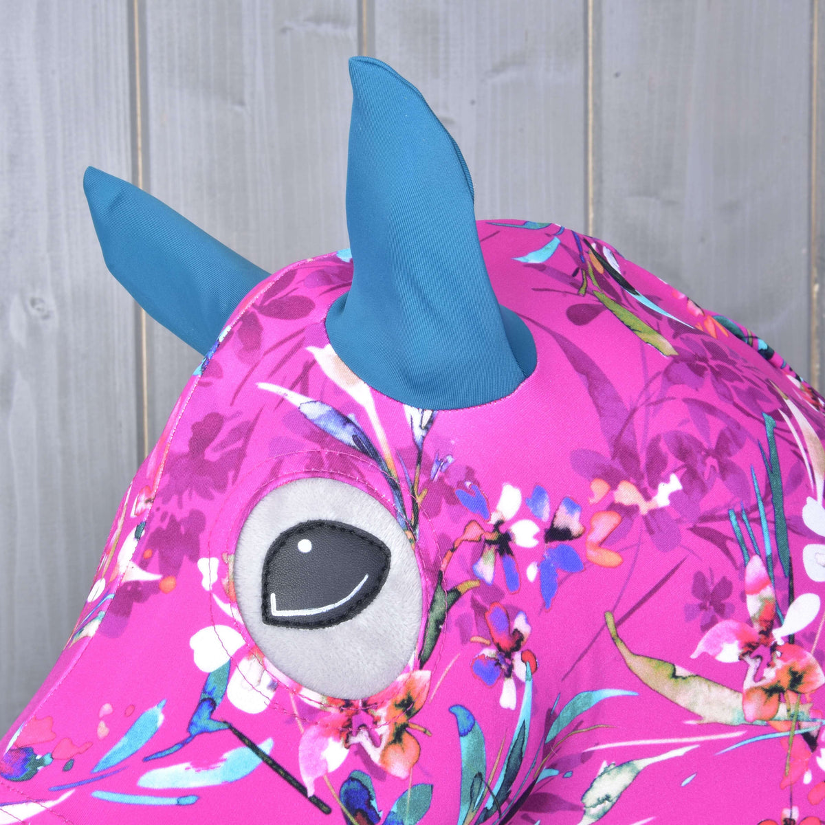 Hobby Horse ochranný oblek Pink Beauty (Lycra) od Hobby Horse LarDen s ružovou kvetovanou látkou, modrými lycrovými ušami/rohmi a veľkým sivo-čierne vyšívaným okom na svetlom drevenom pozadí.