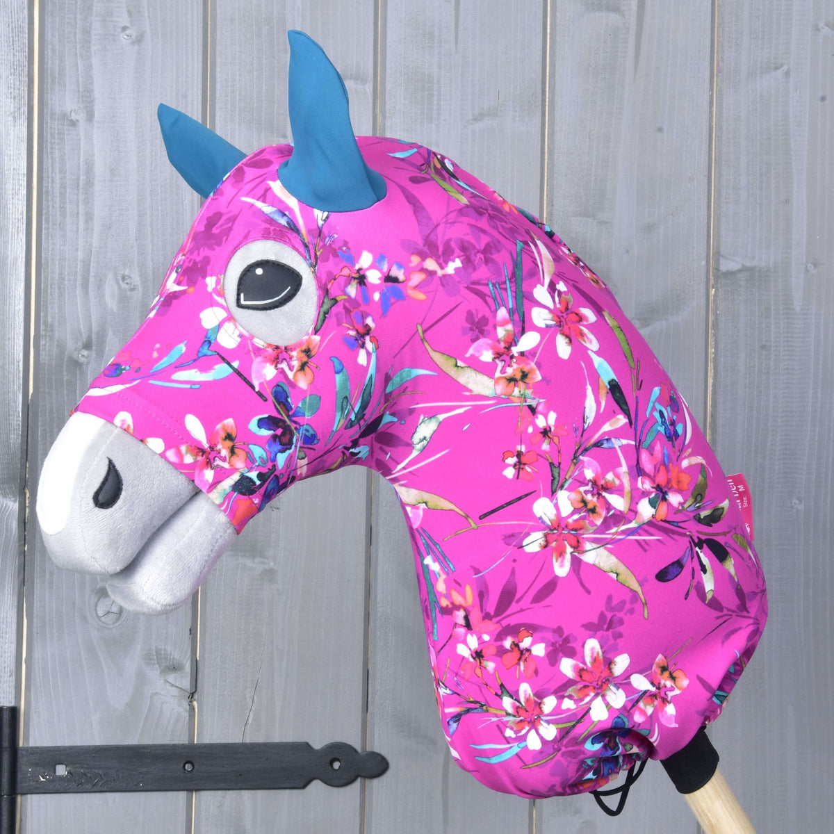 Hobby Horse ochranný oblek Pink Beauty (Lycra) od Hobby Horse LarDen s výraznou ružovou kvetovanou látkou, modrými ušami a bielym ňufákom, nasadený na drevenej tyči pred sivou drevenou stenou.