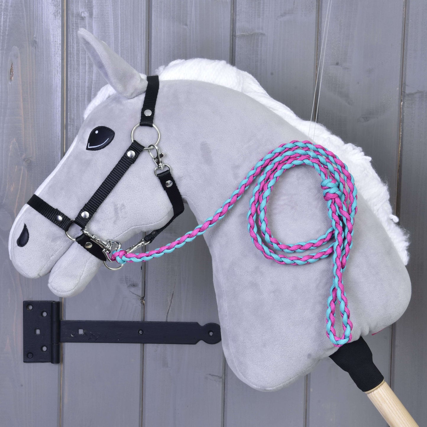 Hobby Horse - pletené vodidlo Pinkie-Blue od Hobby Horse LarDen na sivom plyšovom Hobby Horse s čiernou uzdečkou, bielou hrivou a pleteným ružovo-tyrkysovým vodidlom, zavesenom na drevenej stene. Tyč je súčasťou.