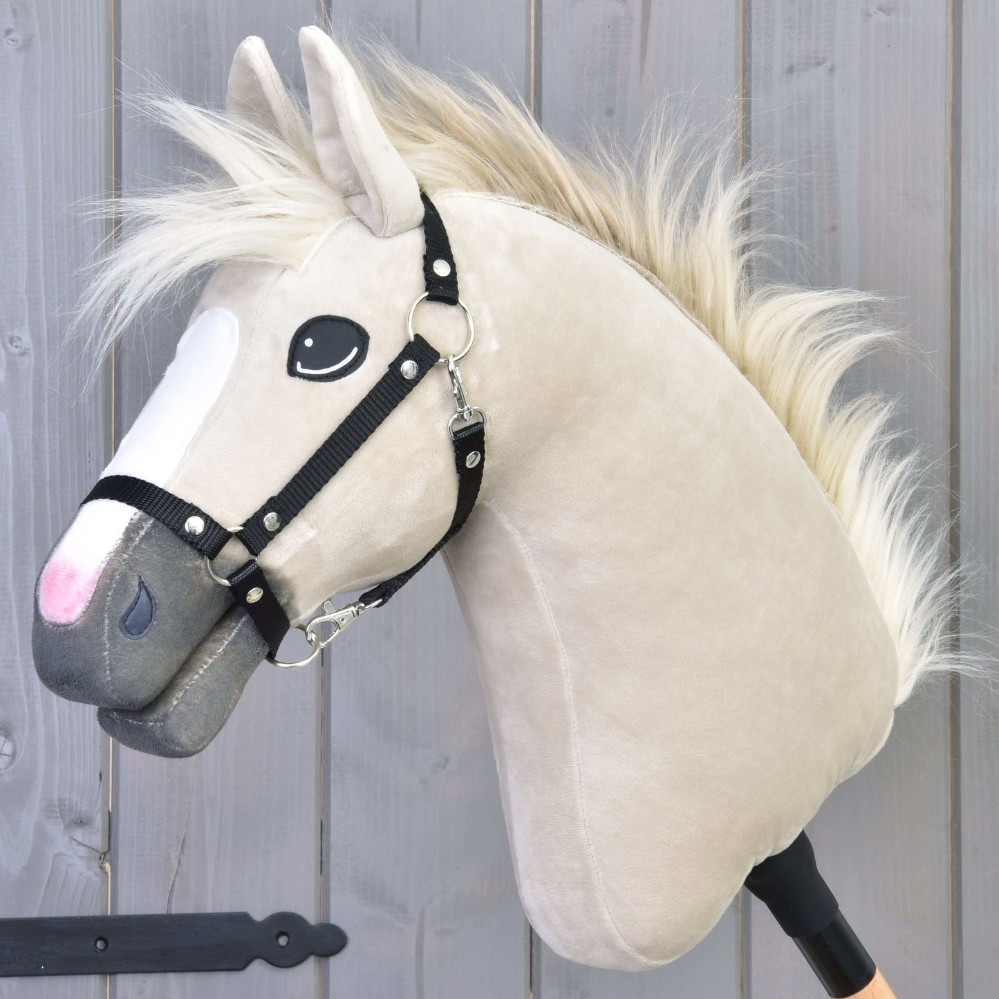 Hobby Horse Pixie s čiernou ohlávkou od Hobby Horse LarDen, s béžovou srsťou, bielou hrivou, čiernou ohlávkou a výraznými očami na tyči, stojí pred sivým dreveným pozadím a odkazuje na legendárny kovový lesk plemena Akhal-Teke.