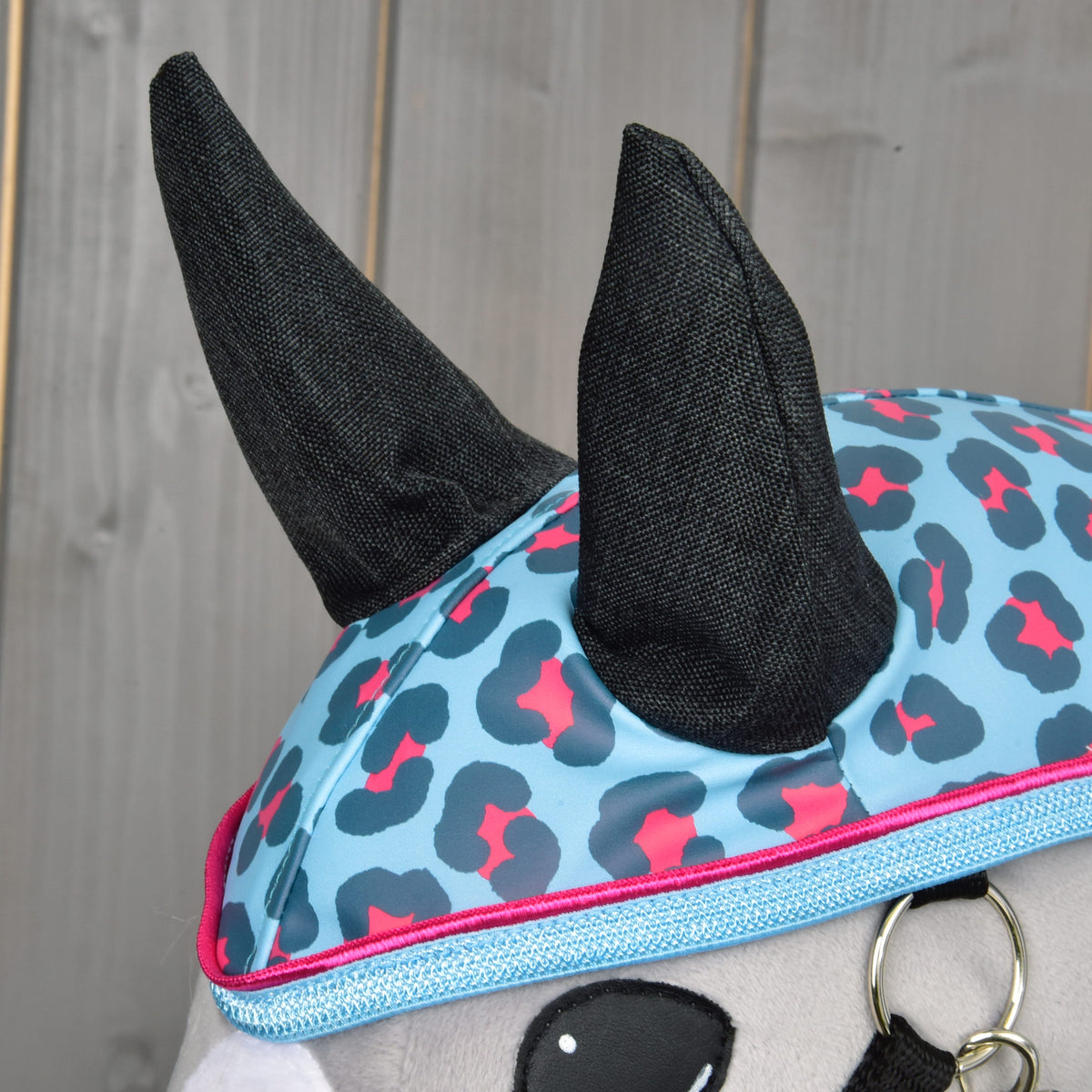 Detail plyšovej hračky Hobby Horse LarDen „Raincoat Blue Leopard“ v modro-ružovej čiapočke s leopardím vzorom a čiernymi rohmi, pripravenej na dobrodružstvá v akomkoľvek počasí, na drevenom pozadí.
