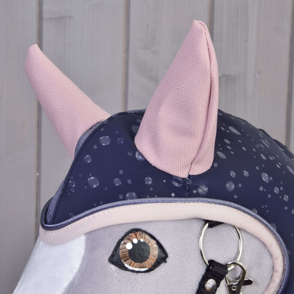 Detail Hobby Horse LarDen Pláštenka Pinkie Blue (softshell): hlava plyšového koňa s veľkým vyšívaným okom, dvoma ružovými ušami, modrou jazdeckou čiapkou s bodkami a uzdečkou s kovovými krúžkami. Pozadie zo svetlých drevených panelov.