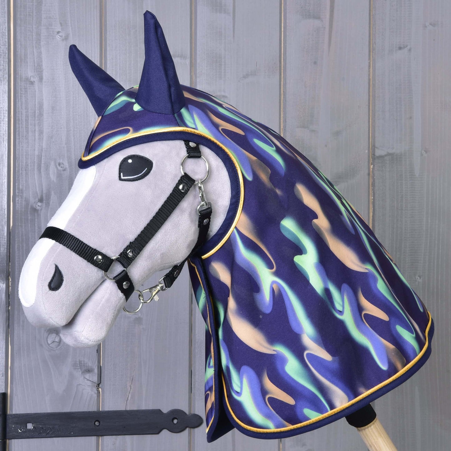 Plyšový Hobby Horse s čiernou uzdečkou má na sebe Pláštenku Rainbow od Hobby Horse LarDen s lesklým viacfarebným dizajnom cez hlavu a krk, na drevenom pozadí.