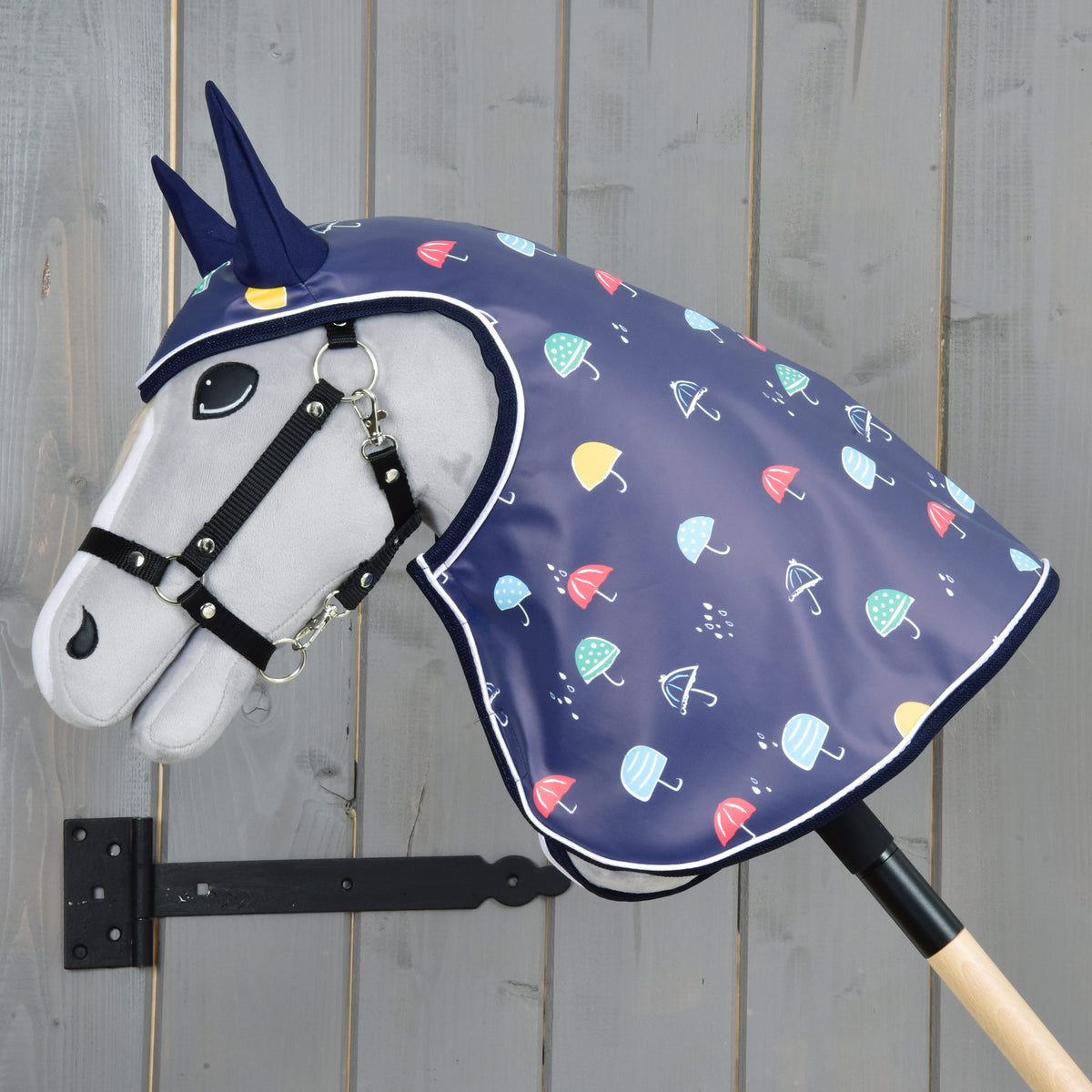 Hobby Horse LarDen „Pláštenka Rainy“ zobrazuje plyšového koňa s uzdečkou a modrým jednorožčím rohom, ktorý má na sebe tmavomodrú pláštenku s farebnými dáždnikmi, upevneného na drevenej tyči na pozadí sivého dreva – ideálny na hru za každého počasia.
