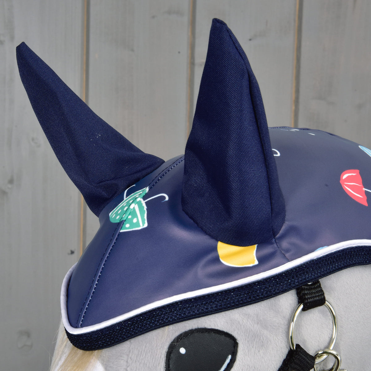 Detail čabraky Hobby Horse LarDen Pláštenka Rainy v modrej farbe so vzpriamenými ušami a výraznými potlačami dáždnikov a dažďových kvapiek – ideálny do každého počasia. Na pozadí svetlosivej drevenej steny.